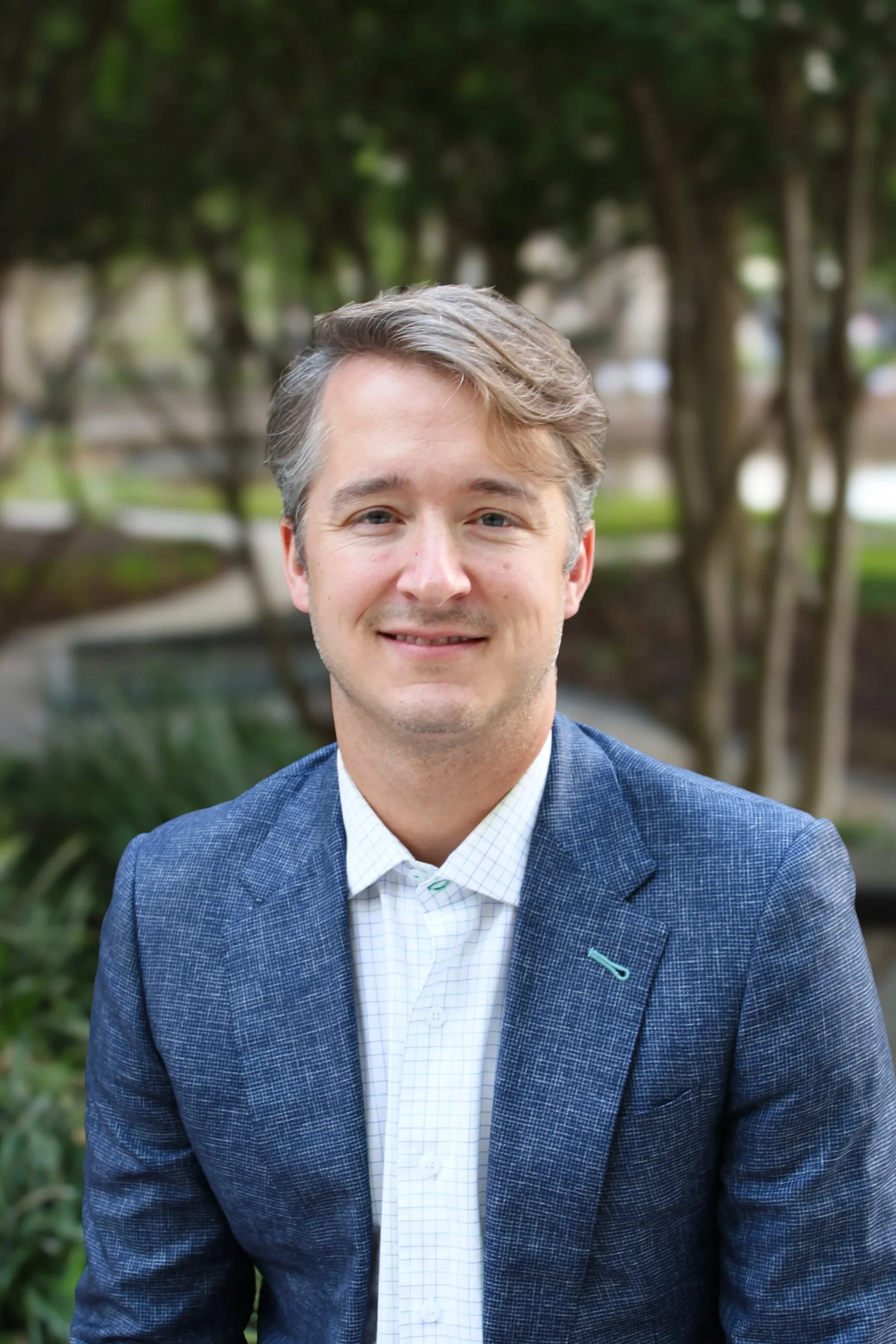 Smiling young man with brown hair, wearing a gray blazer and blue striped shirt, standing outdoors on a city sidewalk with brick buildings and large windows in the background.
