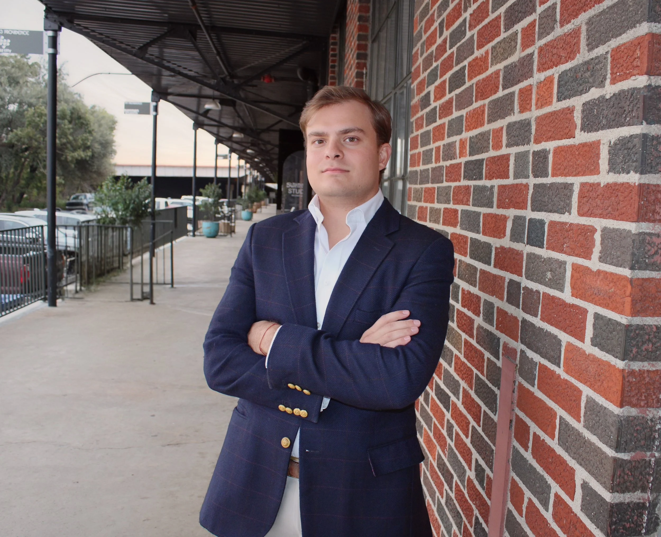 A young man in a navy blazer and white shirt standing with arms crossed against a brick wall outside a building at sunset.