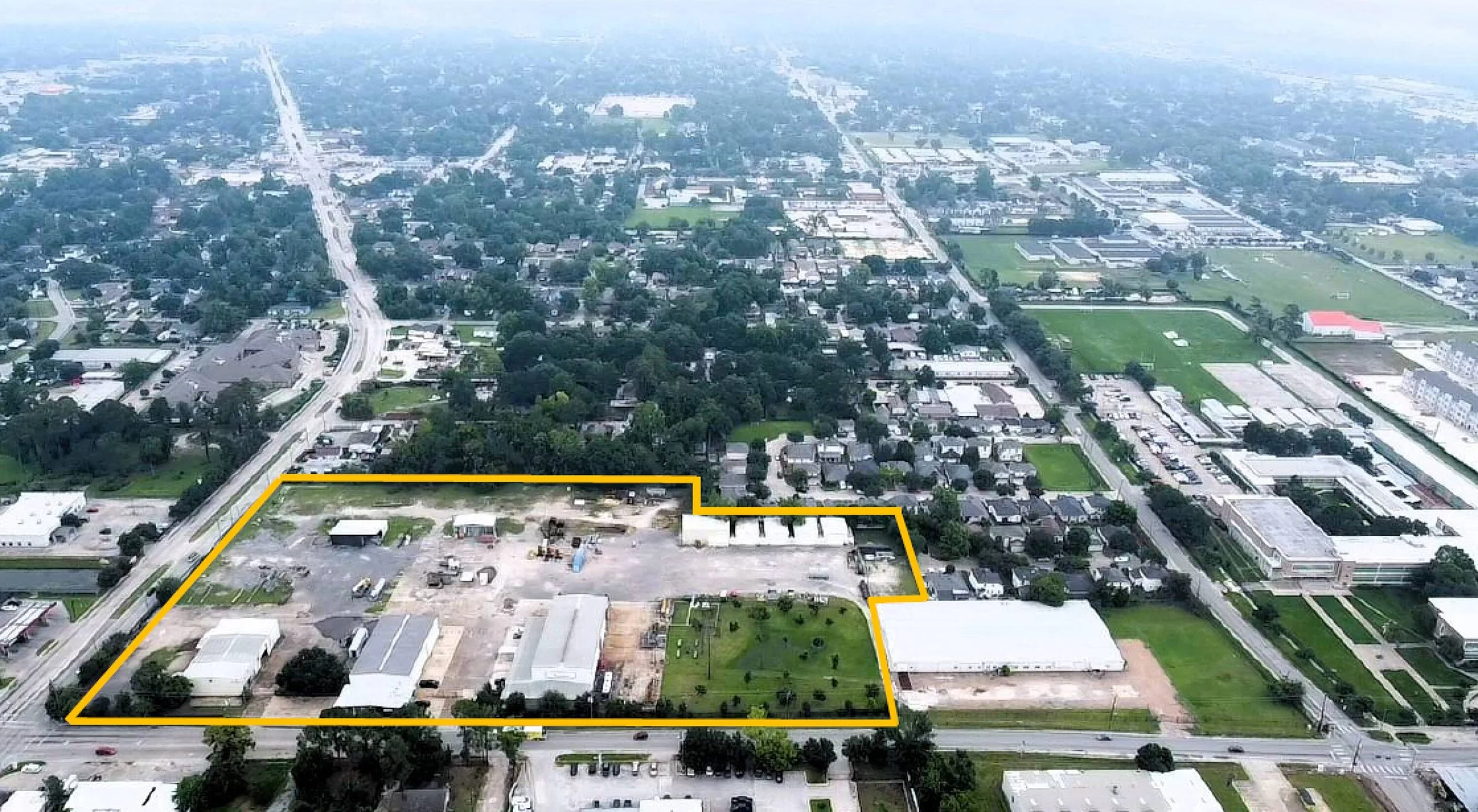 Aerial view of a city neighborhood with a large outlined vacant lot featuring some buildings, green space, and parking areas.