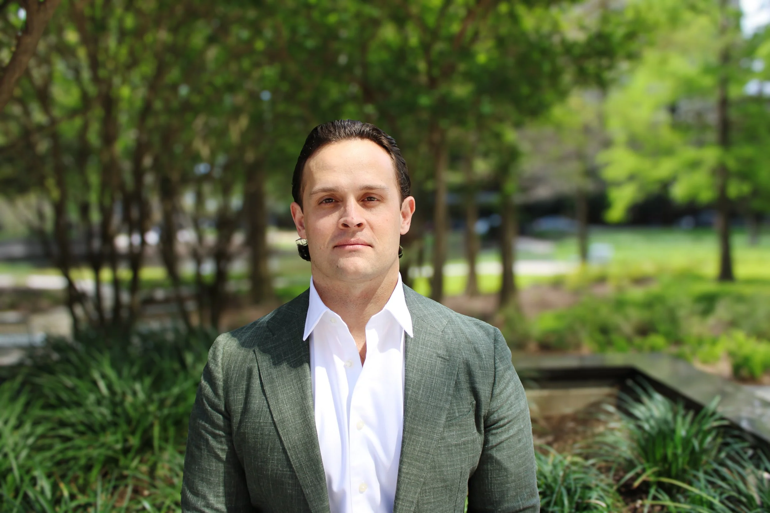 Man in a dark green blazer and beige pants standing against a brick wall outside a building, looking at the camera with a serious expression.