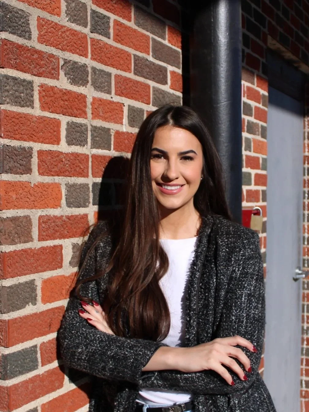 A young woman with long brown hair wearing a black and white textured blazer, white shirt, and light blue jeans, standing with arms crossed in an indoor space with metal panel walls and wooden furniture.