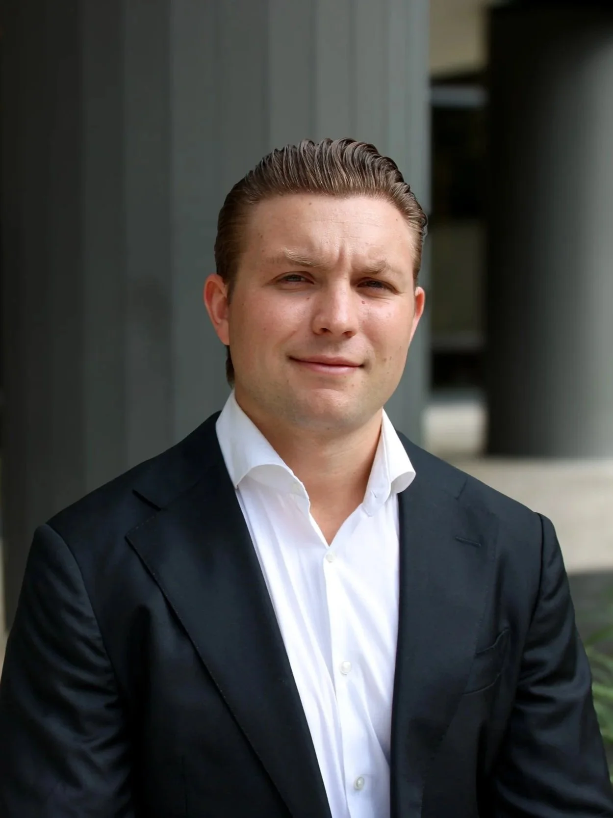 Young man in a suit standing outside under a covered walkway next to a brick building, with potted plants and benches visible in the background.