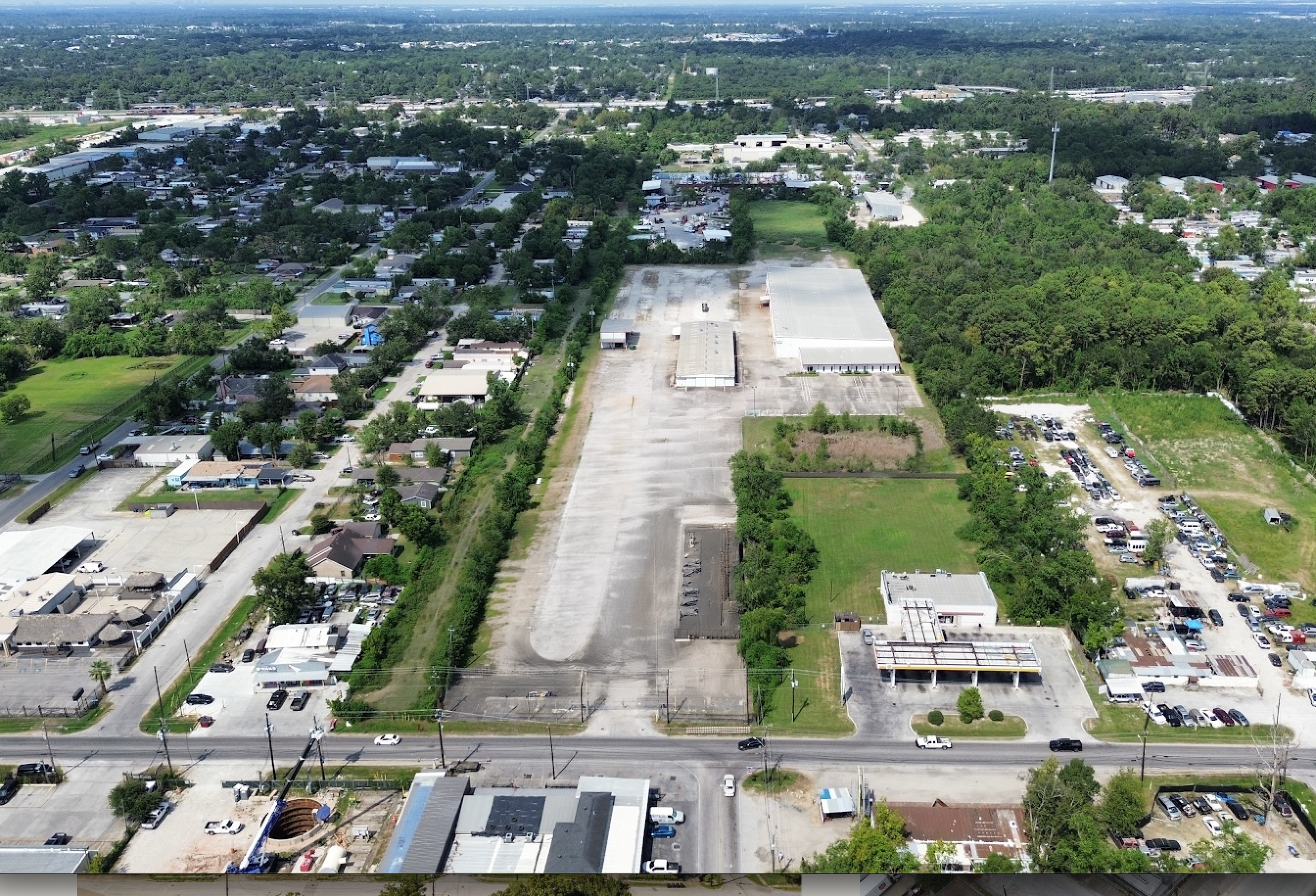 Aerial view of an urban area with residential homes, commercial buildings, parking lots, and green spaces, along with an under-construction or demolished plot and surrounding streets.