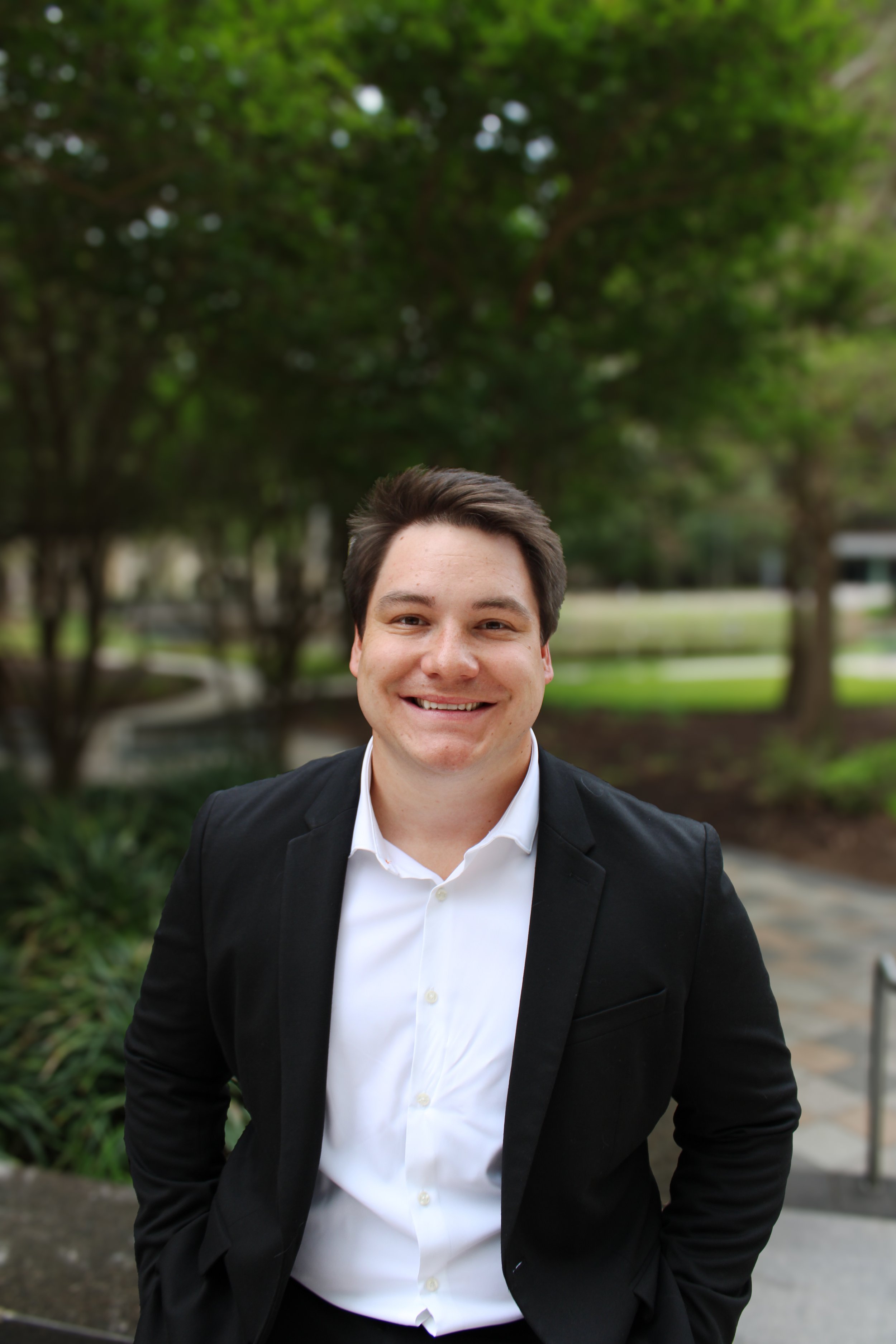 A young man in a business suit standing indoors near colorful framed posters, with a potted tree in the background.