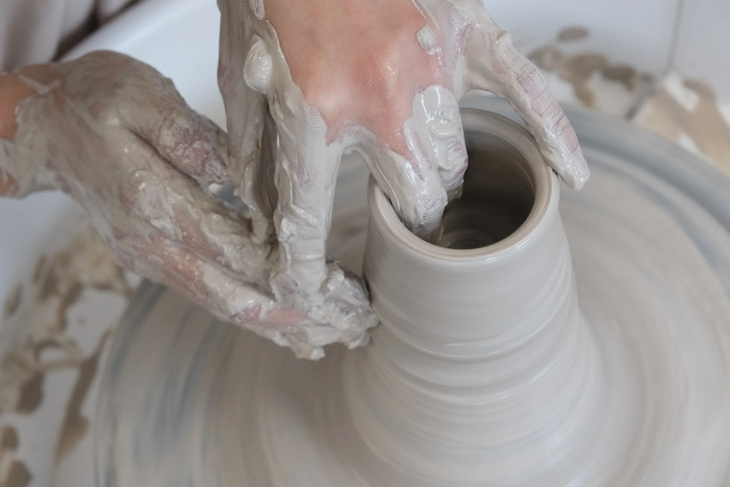 Hands covered in wet clay shaping a pottery vessel on a wheel.