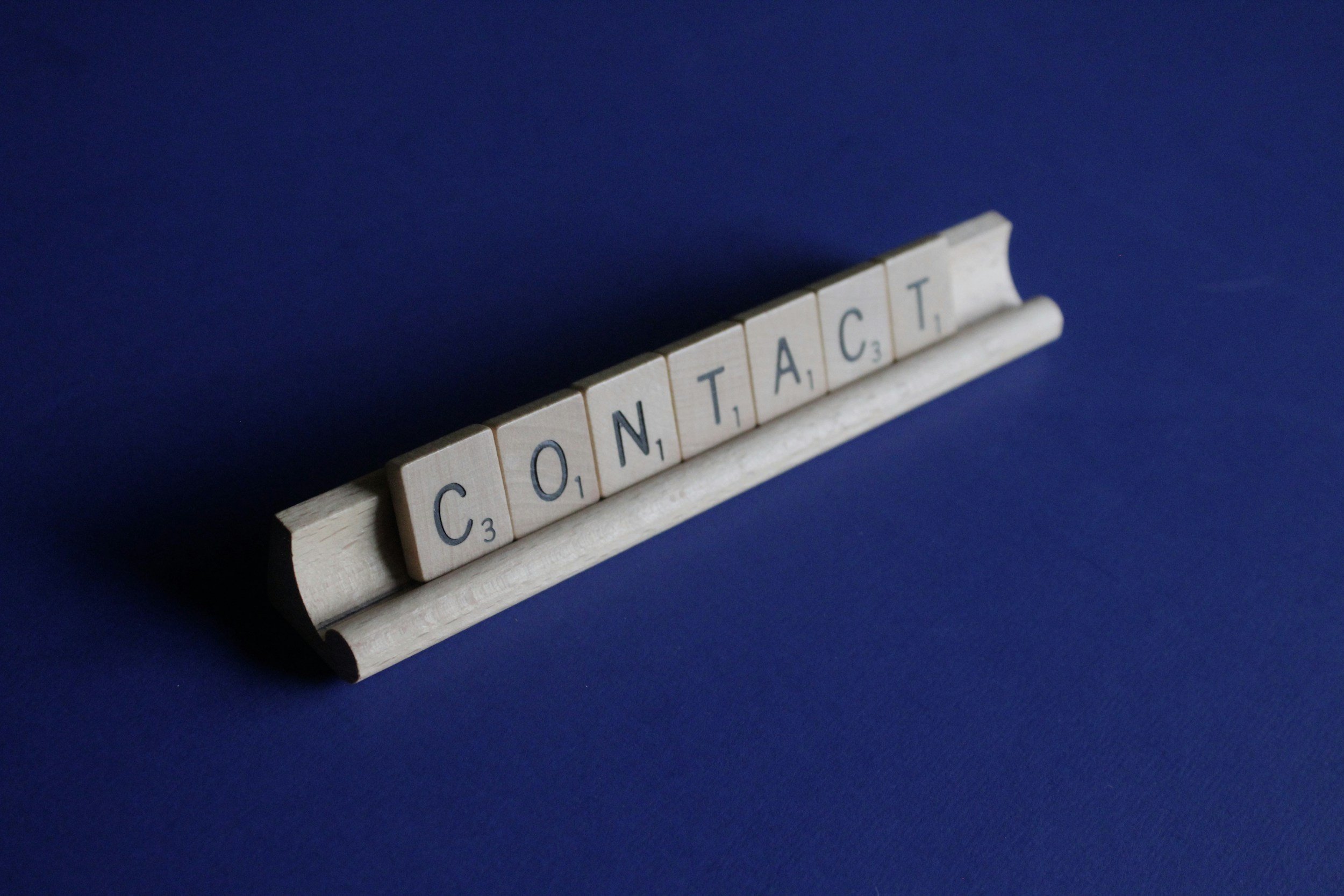 Scrabble tiles spelling 'CONTACT' on a wooden rack against a blue background.