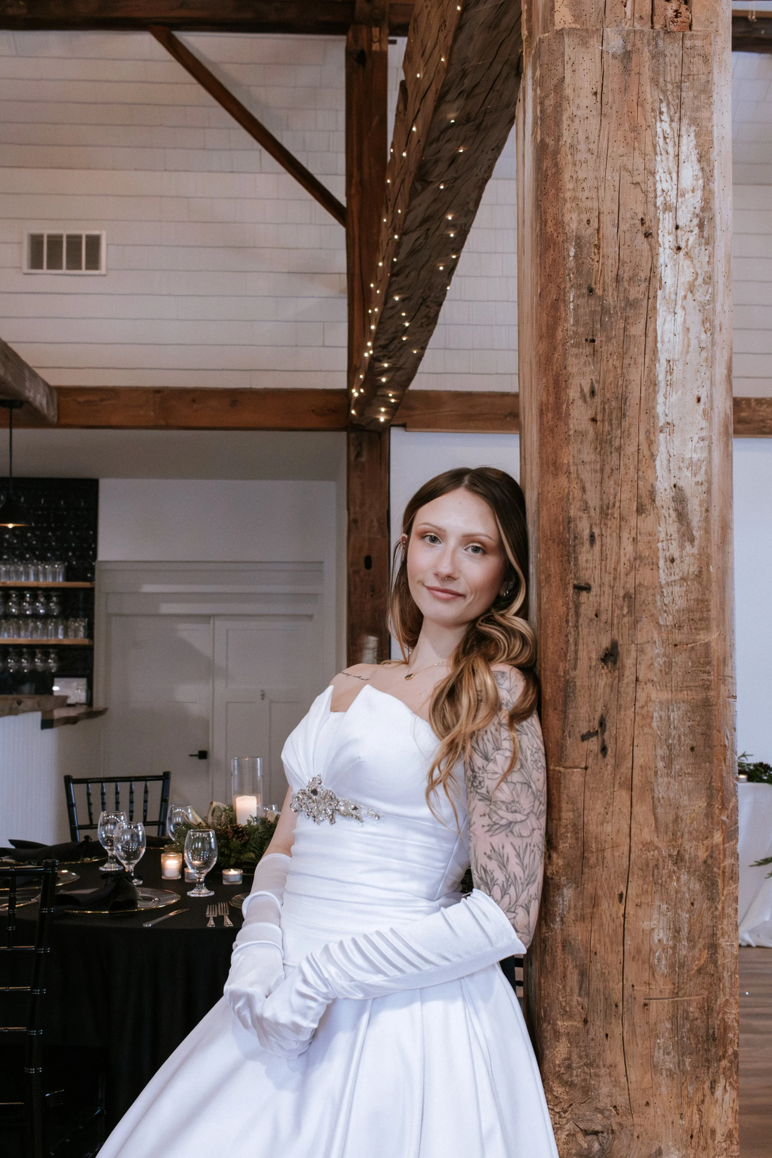A bride in a white wedding dress with long gloves, leaning against a wooden column in a decorated wedding venue.