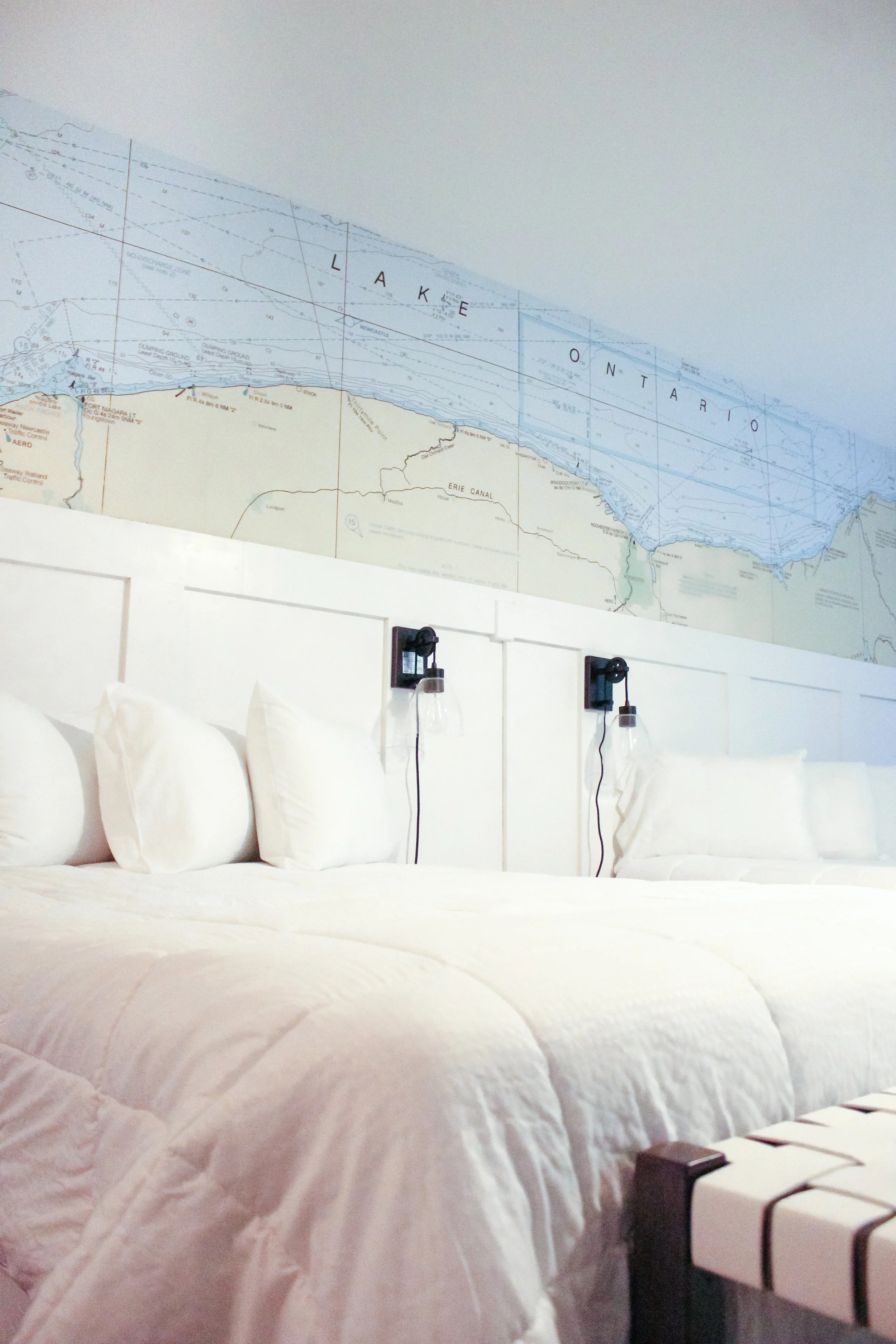 Bedroom with white bedding and a map of Lake Ontario on the wall above the bed, featuring two wall-mounted black sconces and a white cushioned bench at the foot of the bed.