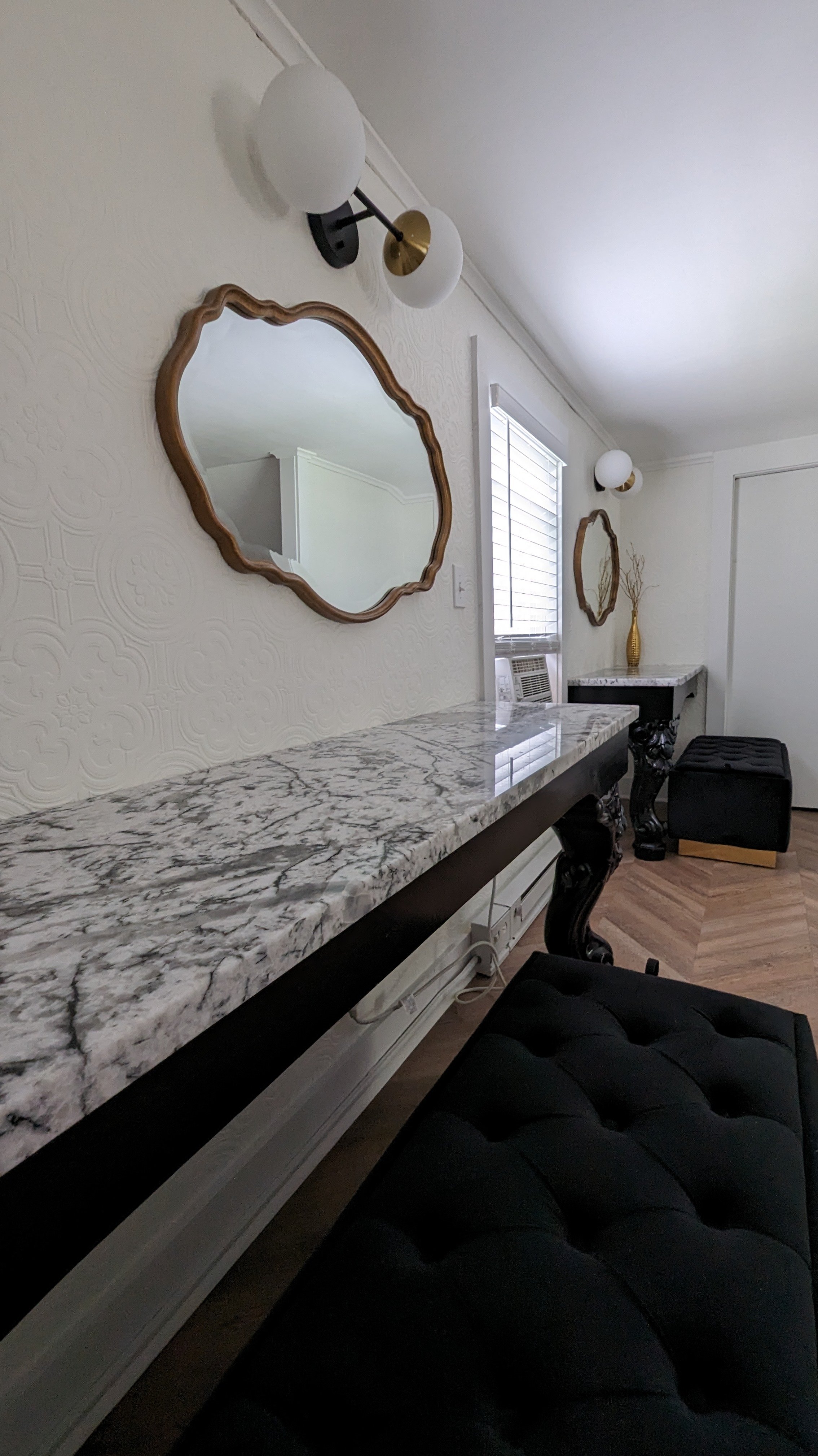 Interior of a room with a marble-topped console table, ornate mirror, and tufted bench. The room features wall sconces and decorative elements.