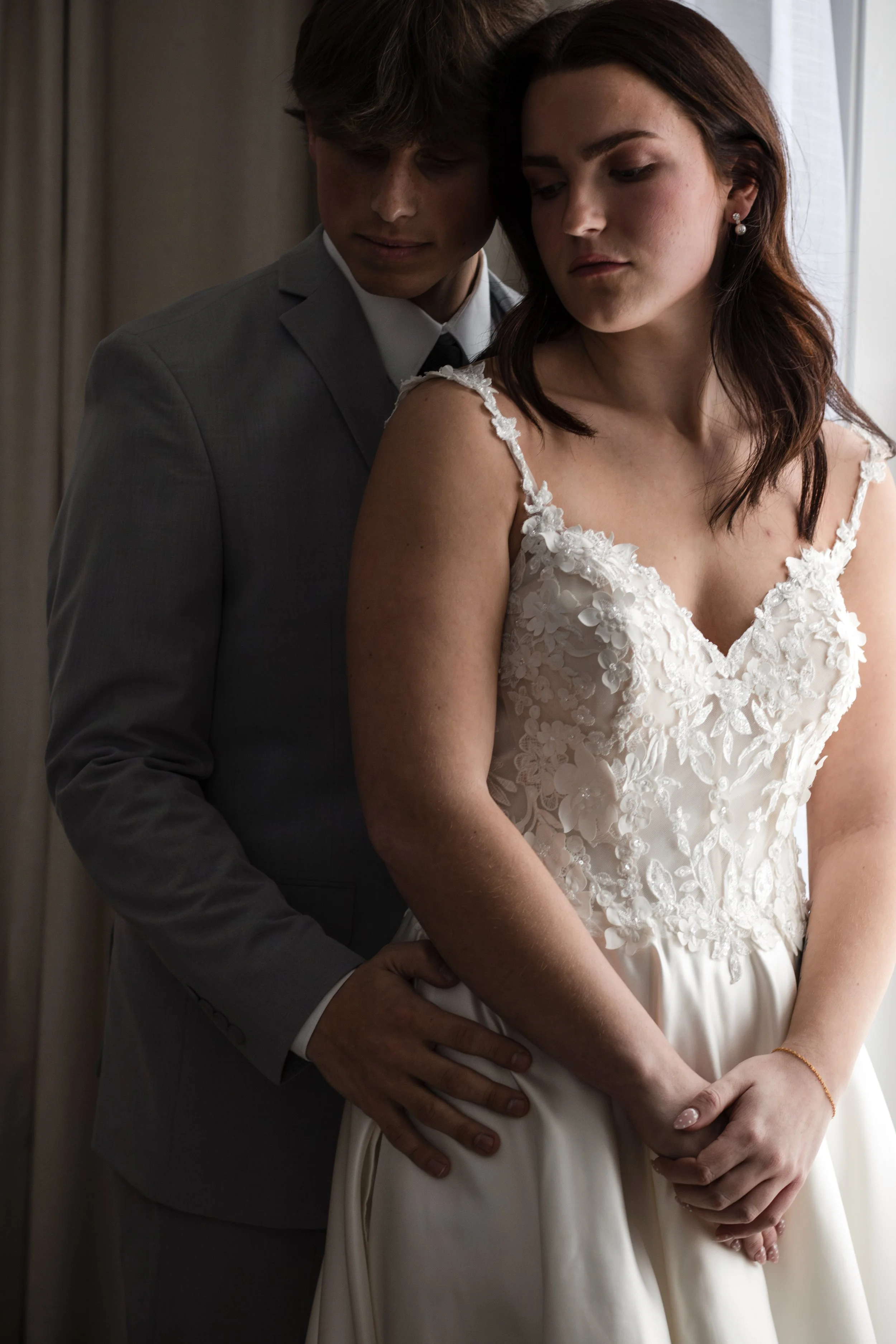 A bride and groom share an intimate moment, with the groom standing behind the bride, gently holding her waist. The bride is wearing a lace wedding dress, and the groom is dressed in a gray suit. They are near a window, with soft natural light illumi