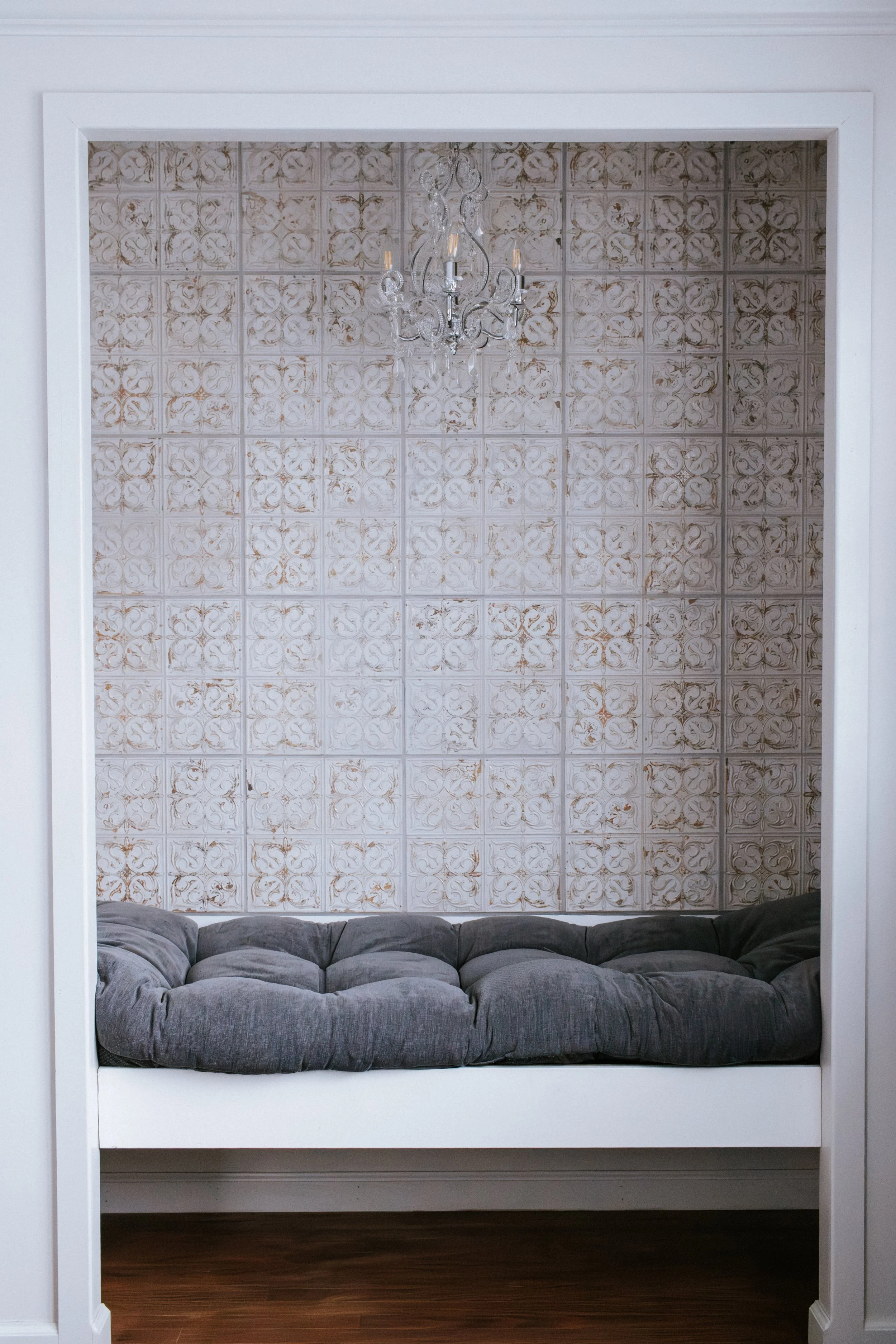 A built-in window seat with plush gray cushions, framed by white walls, a vintage patterned tile wall with a chandelier overhead, and wooden flooring.