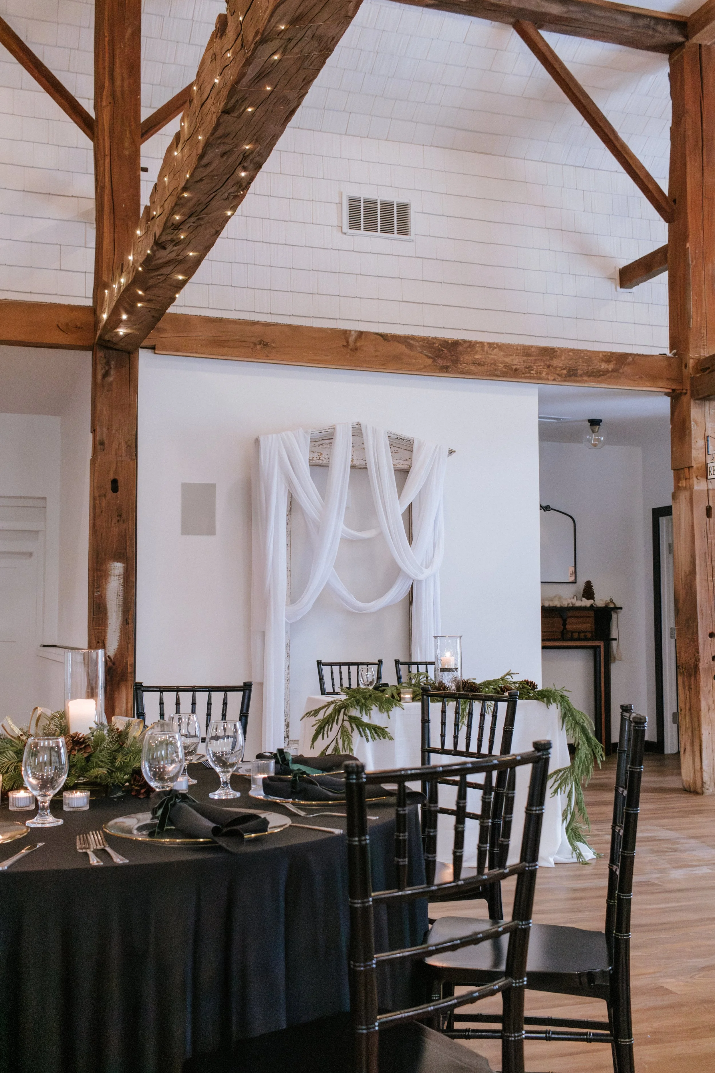 Wedding reception table with black tablecloth, glassware, candles, greenery, and a backdrop of draped white fabric on a wooden frame in a rustic venue with exposed beams.