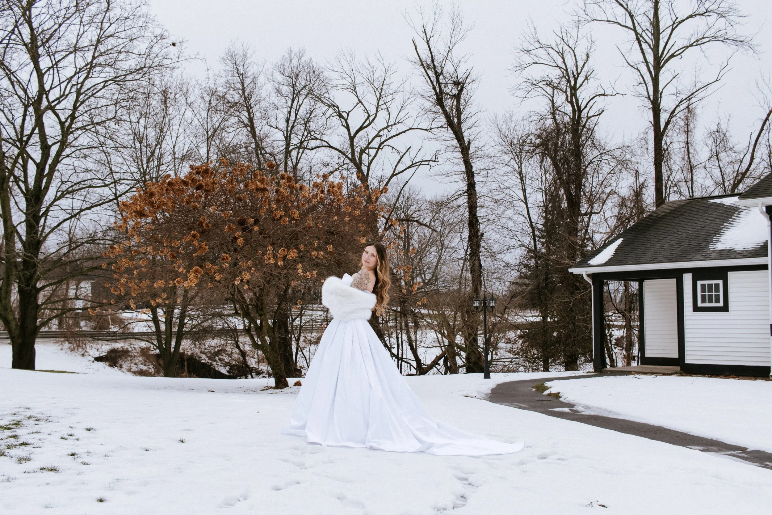 A woman in a white wedding gown and fur stole standing on snow-covered ground outdoors in winter with bare trees and a black-and-white house nearby.