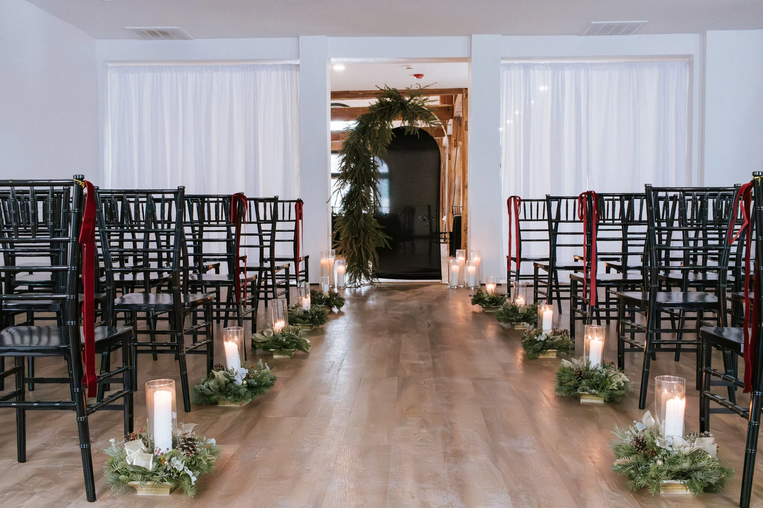 Wedding aisle decorated with candles and greenery leading to an archway with a hanging greenery arrangement, chairs on both sides with red ribbons, white curtains in the background.