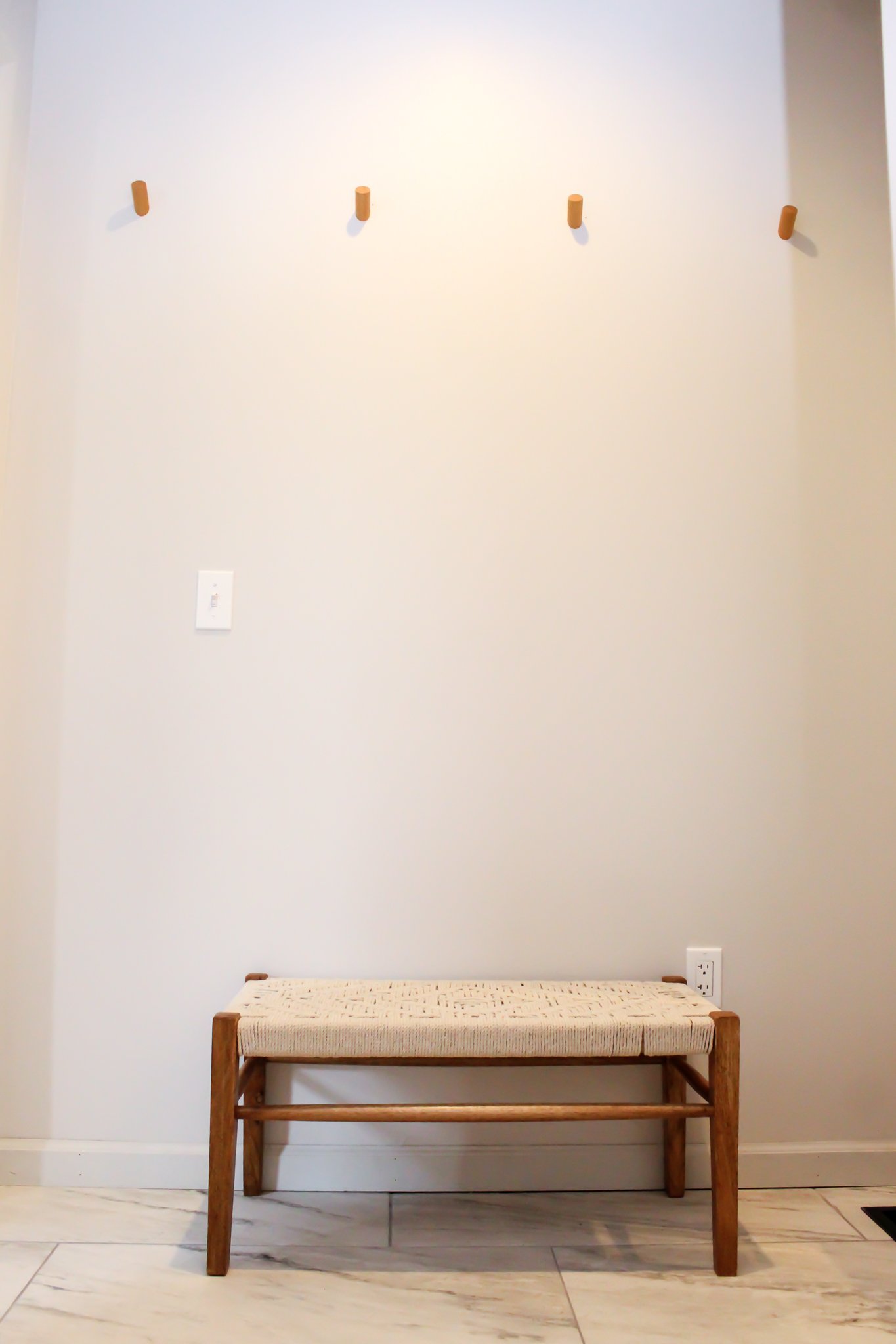 Minimalist entryway with wooden bench and four wall hooks above on a light-colored wall, tiled floor, and a light switch and outlet nearby.