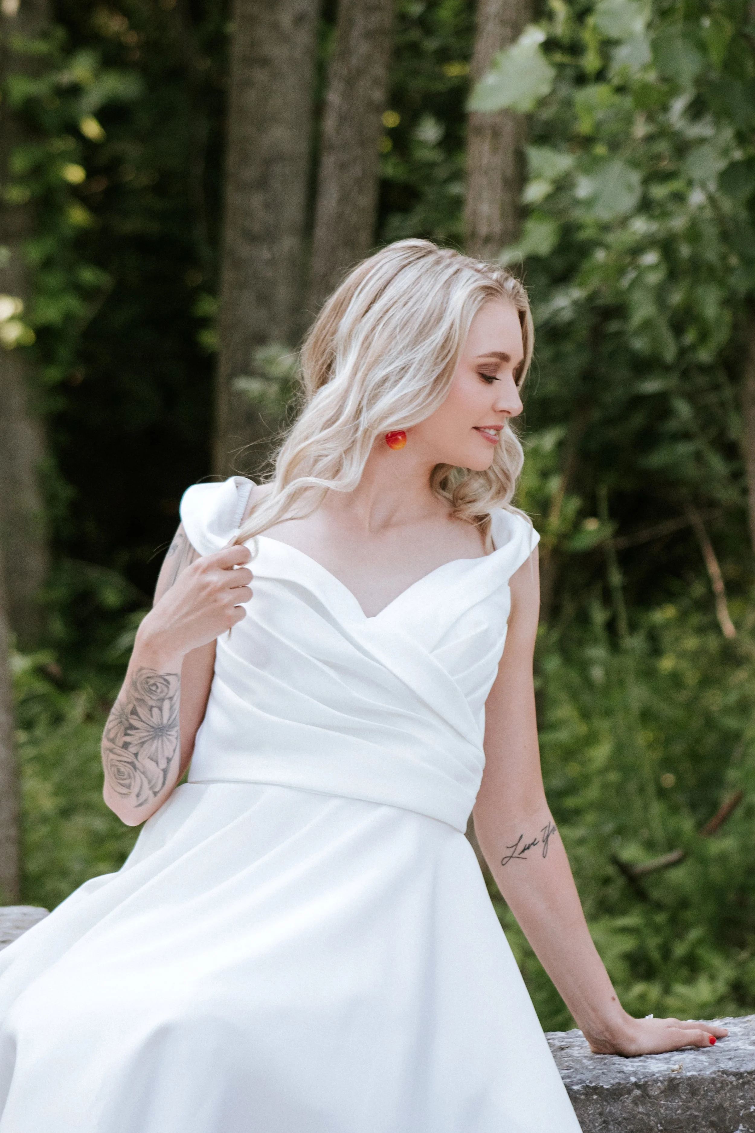 Woman in a white dress with tattoos on her arms, standing outdoors among trees, looking to the side, with blonde hair and red earrings.