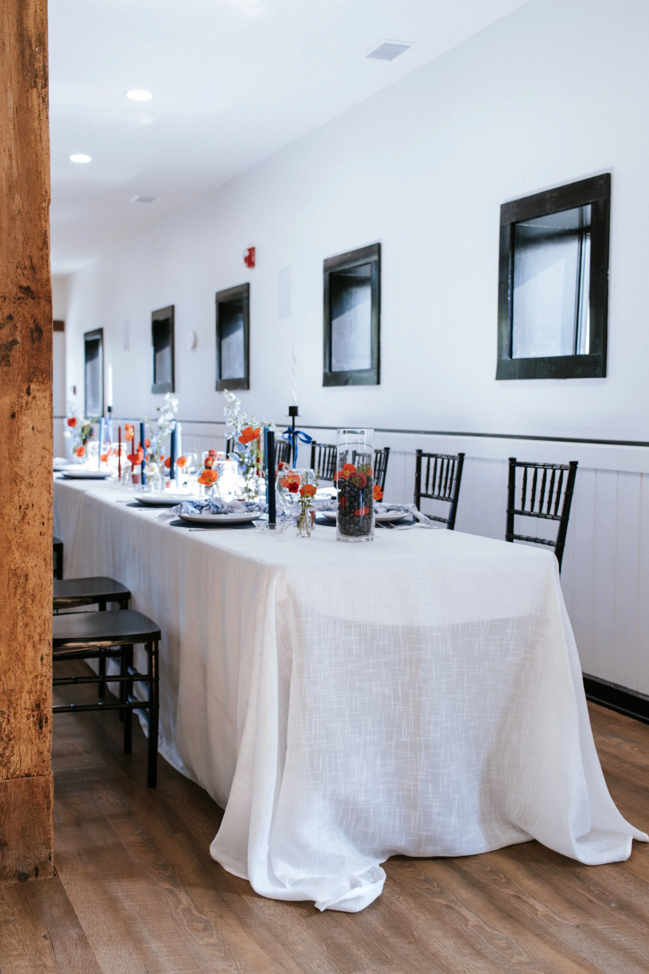 Long dining table covered with white tablecloth, decorated with orange and white flowers, candles, and glass jars, set for a celebration in a bright room with white walls, black framed windows, and wooden flooring.