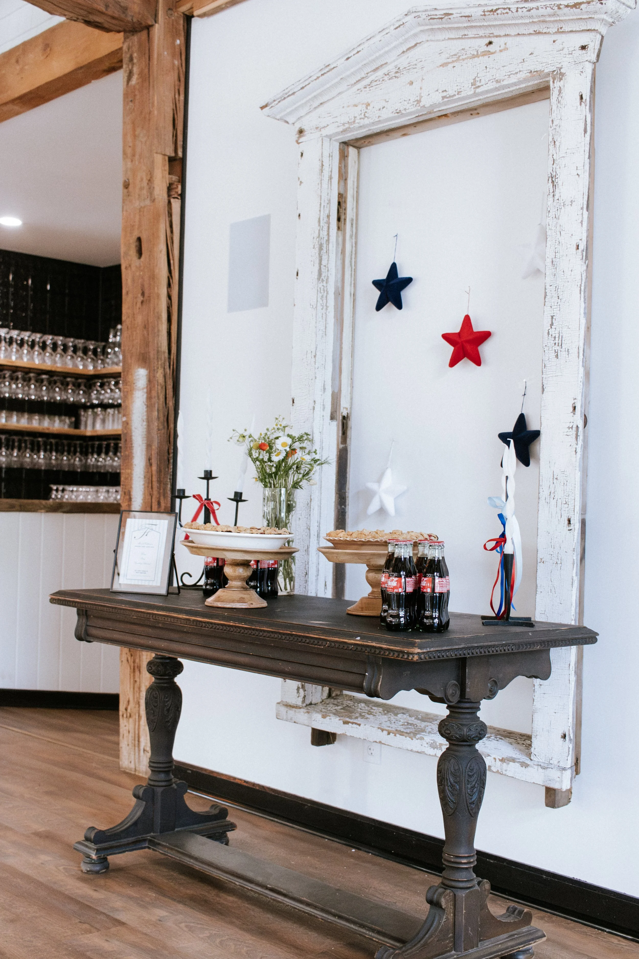 Table with soda bottles, serving trays with snacks, a flower vase, and patriotic decorations in front of a rustic white door with star ornaments.