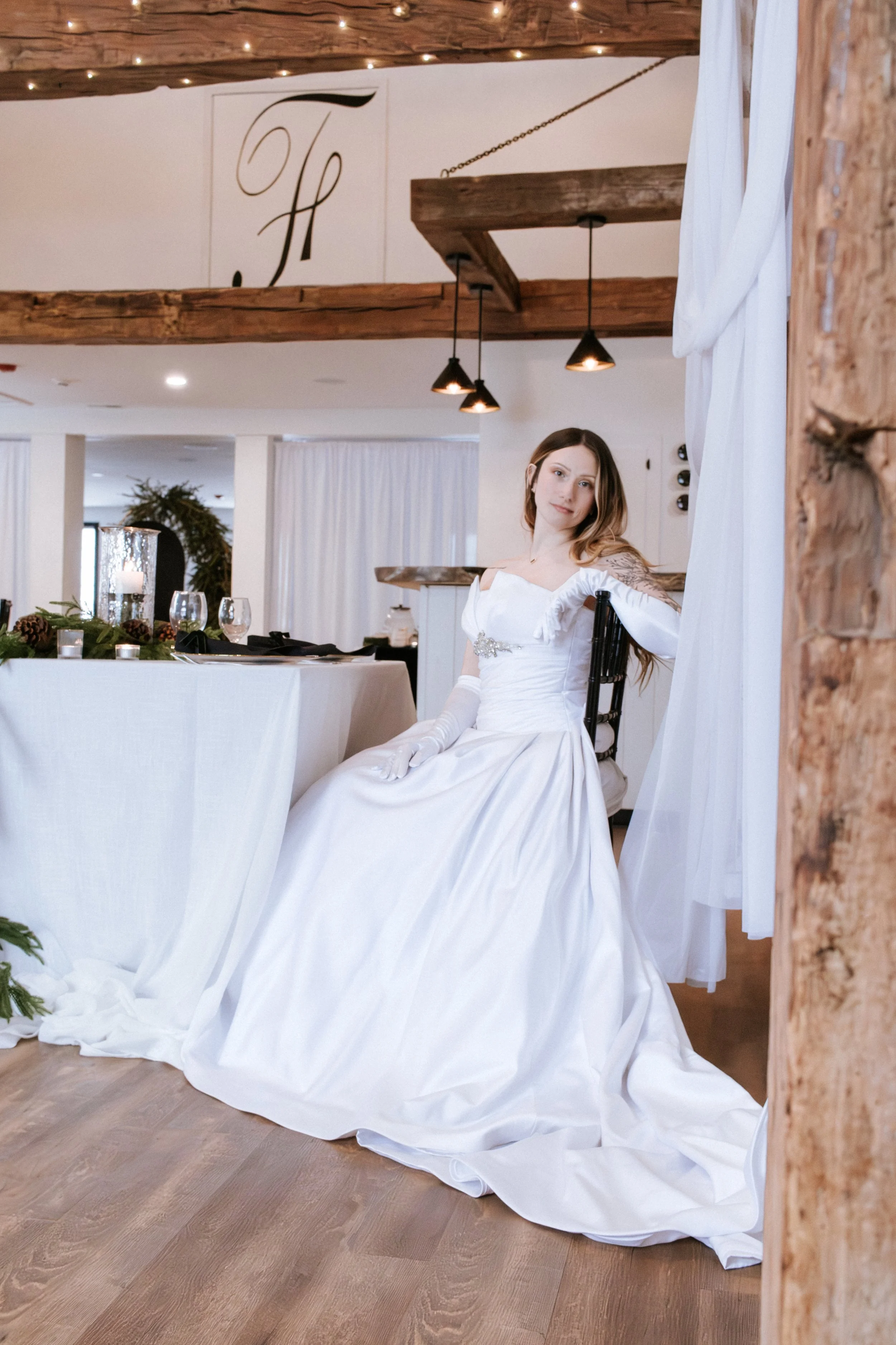 A bride in a white wedding gown sitting at a decorated table in a rustic wedding venue.