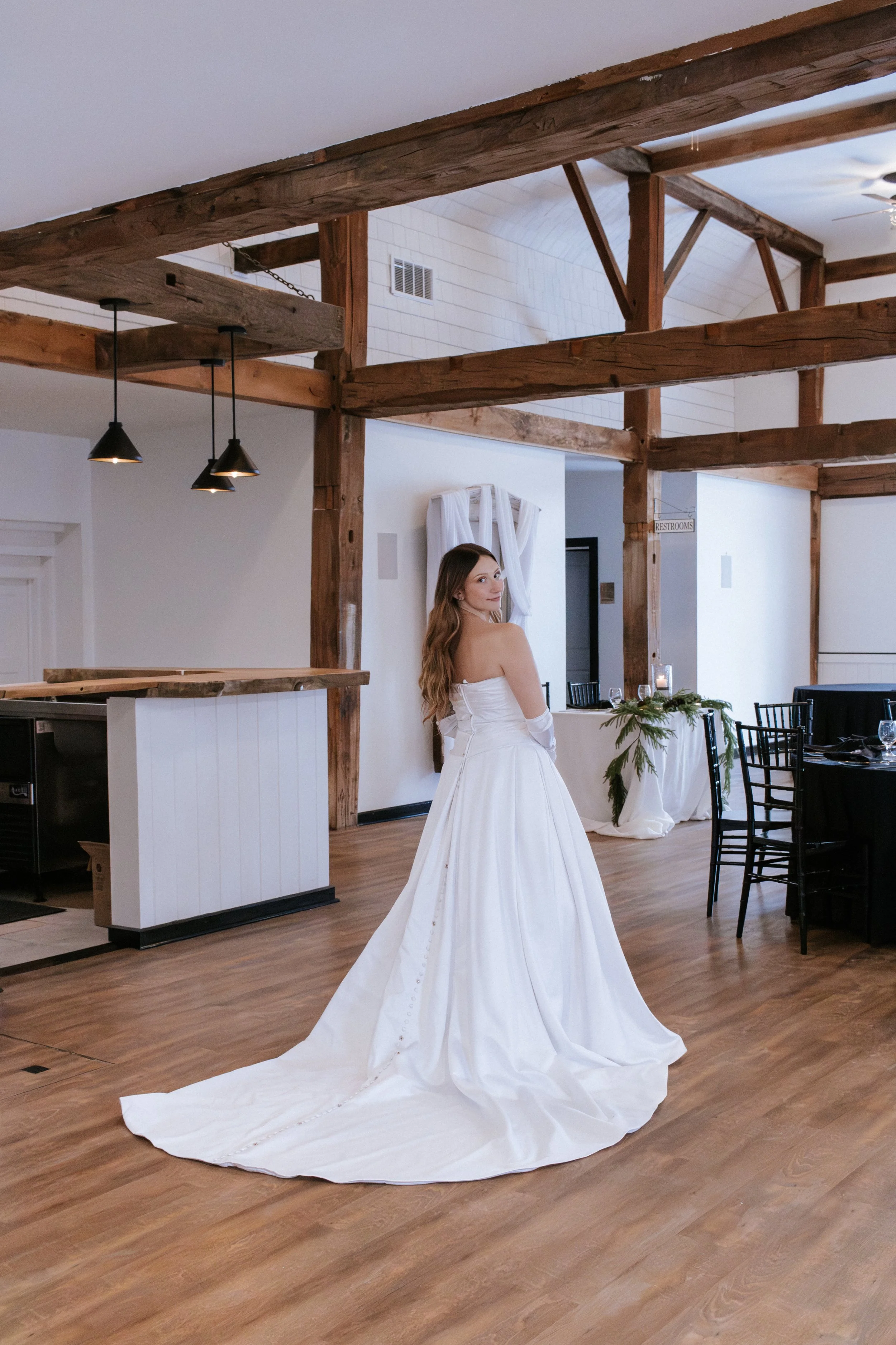 A woman in a white wedding dress with off-shoulder sleeves standing in a rustic indoor venue, with wooden beams and a long train behind her.