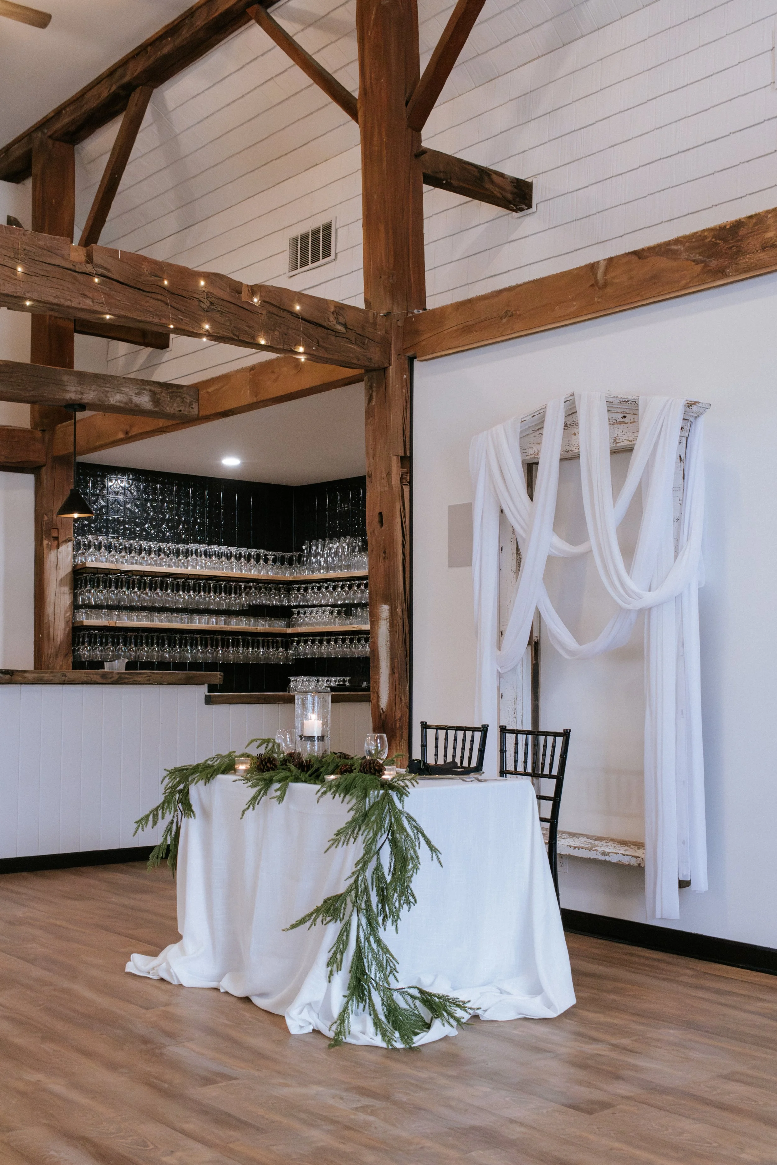 Rustic indoor event space decorated with greenery and white drapery, featuring a table with candles and two black chairs, and a background of hanging glassware and wooden beams.
