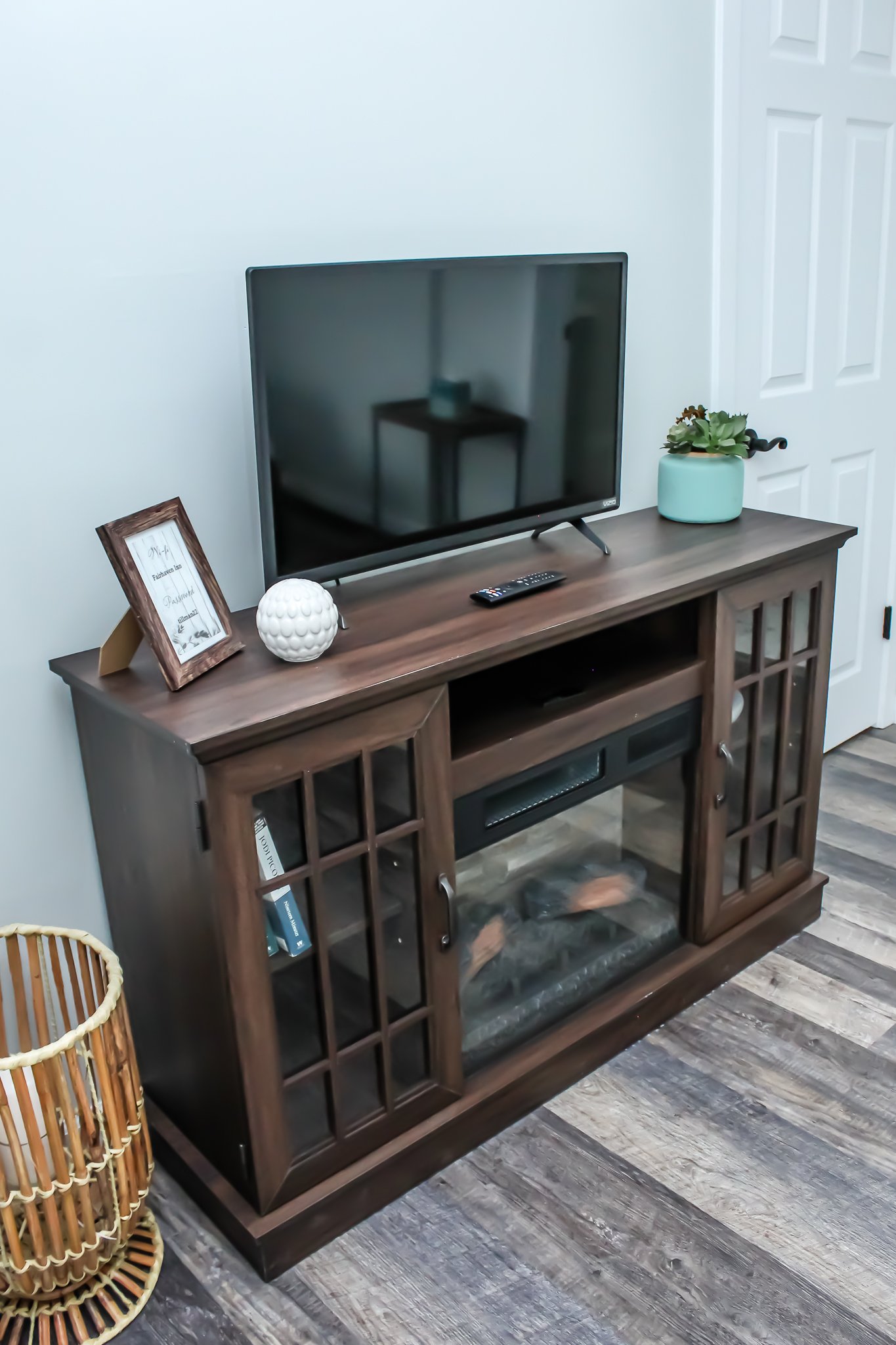 Living room setup with a flat-screen TV on a wooden entertainment stand, decorative plants, picture frame, and remote control, with a basket and wood flooring.