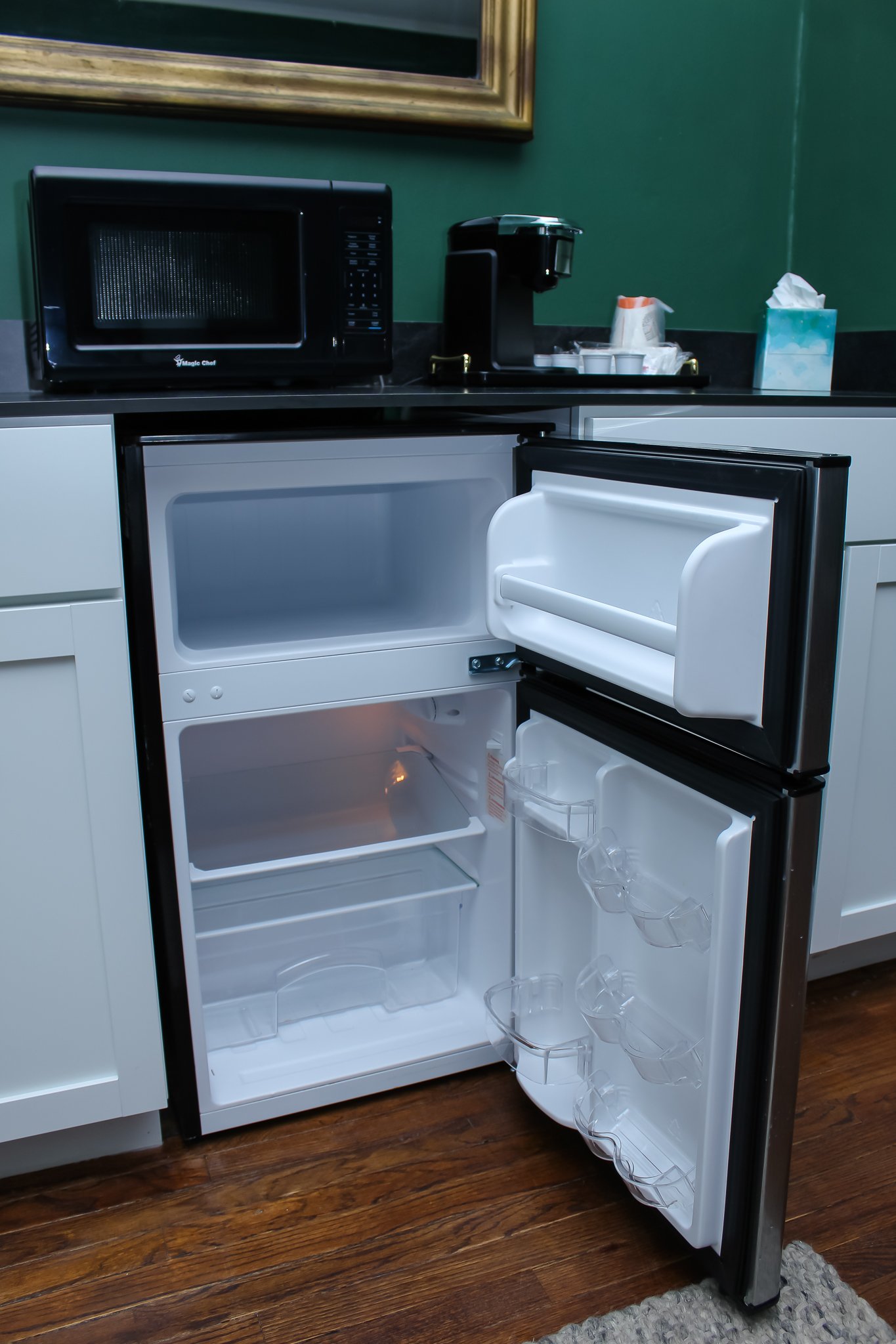 Open mini fridge with empty shelves and door compartments, a microwave on top, coffee maker, tissue box, and cups on a countertop.