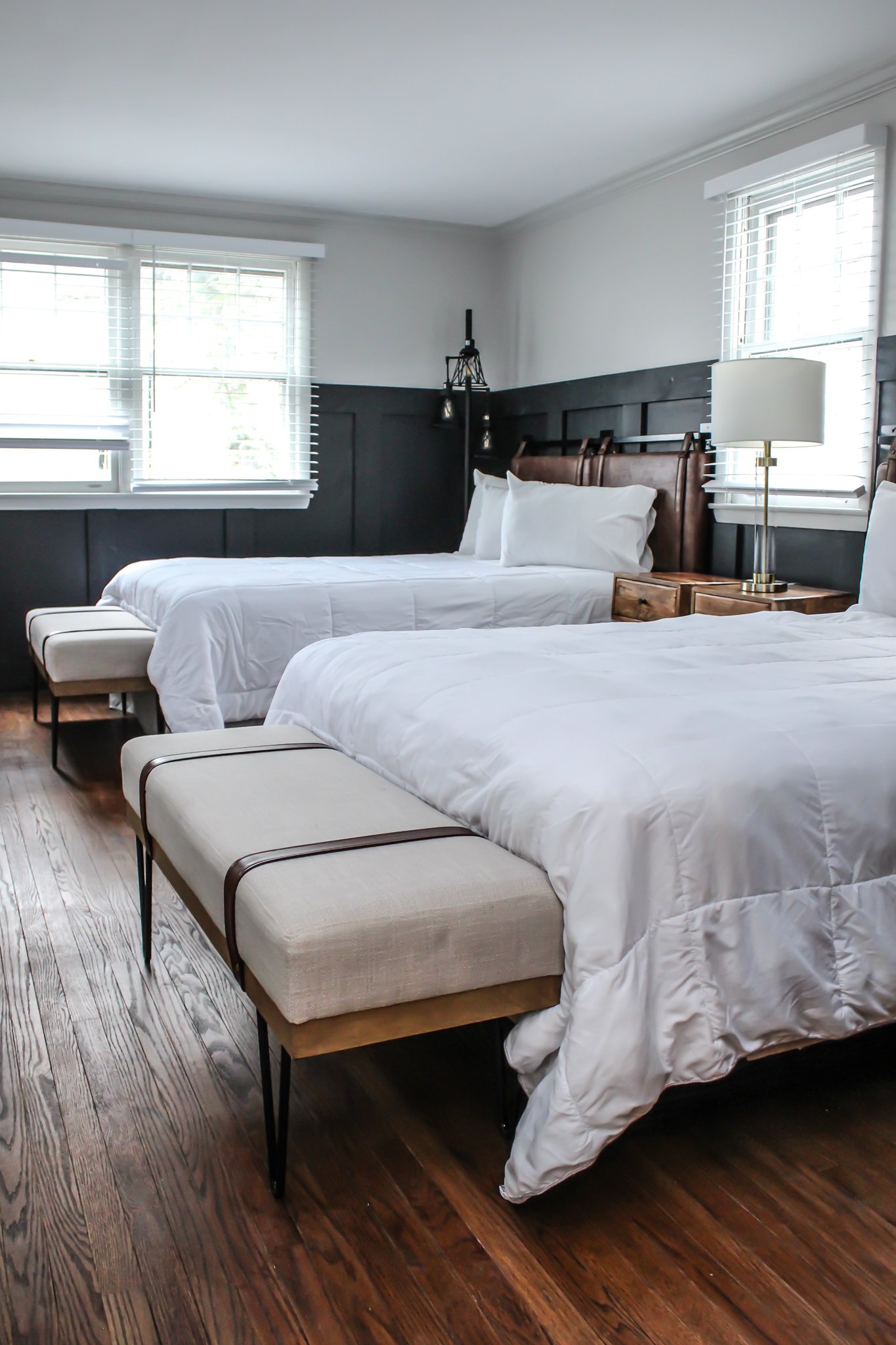 A modern bedroom with two beds, white bedding, and wooden accents. A nightstand with a lamp is placed between the beds. The room has wooden floors and large windows with blinds.