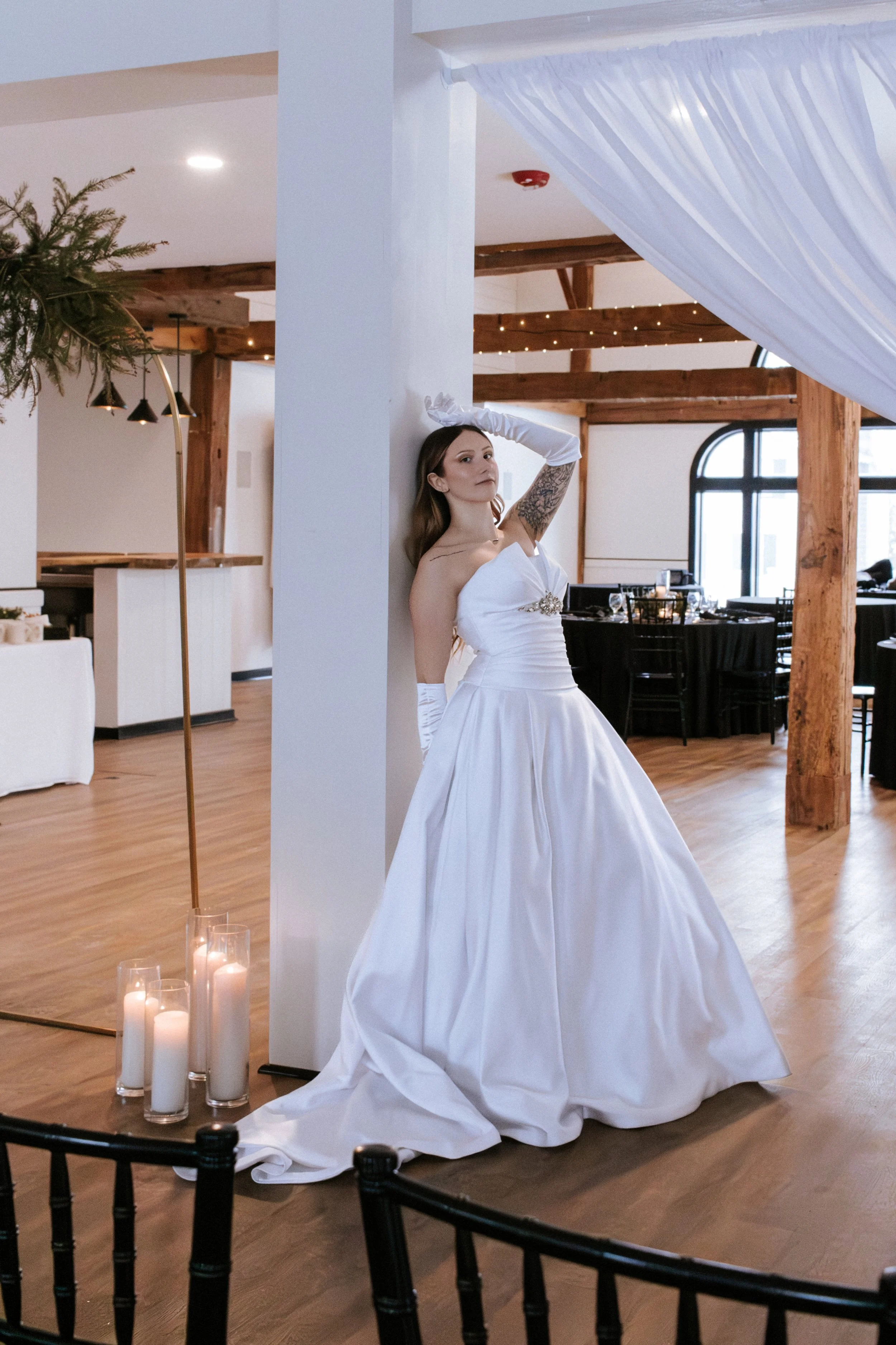 A bride in a white wedding gown and long gloves poses indoors, leaning against a white column, with candles on the floor nearby, in a decorated venue with wood beams and large windows.