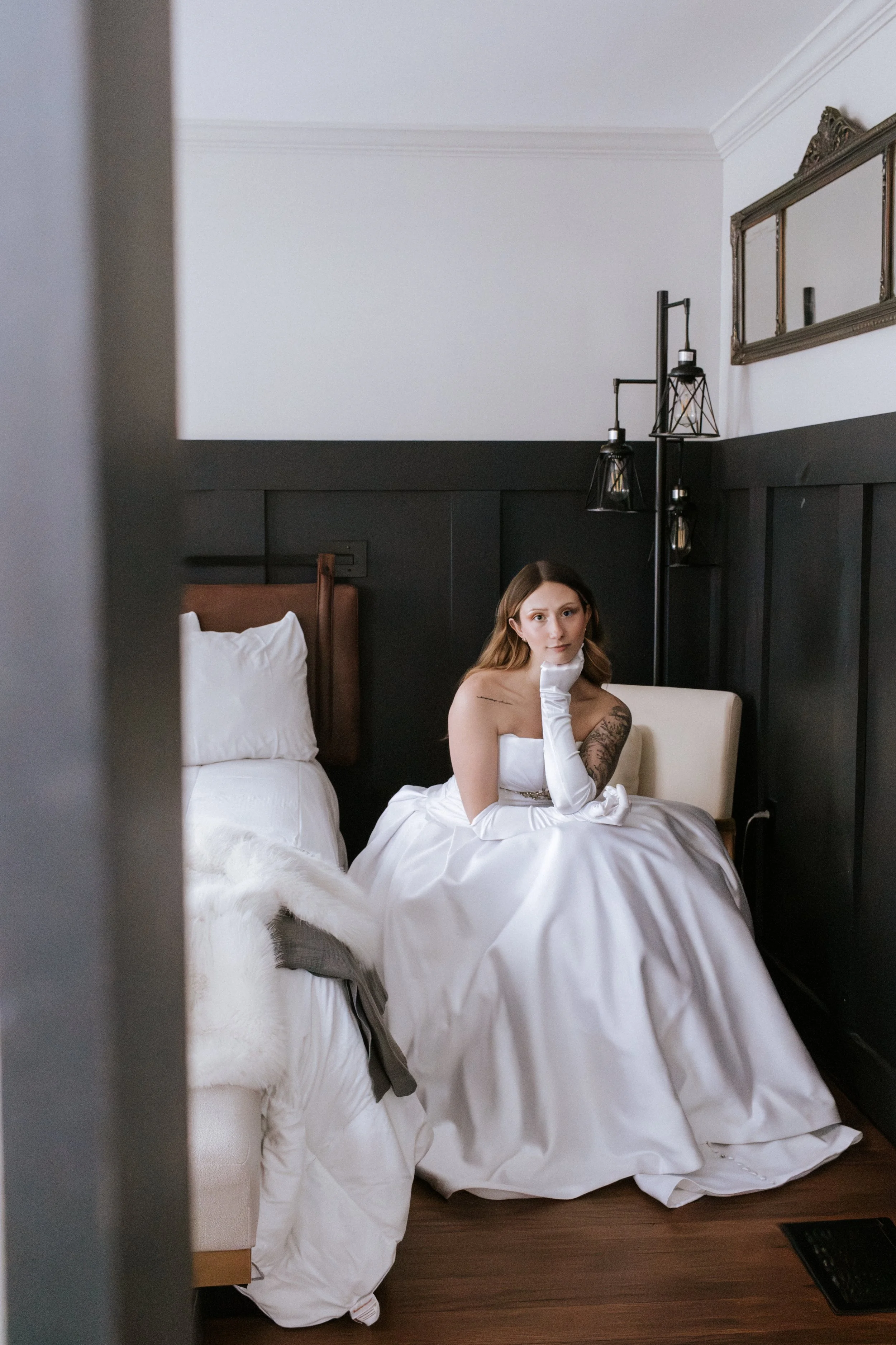 A woman in a white wedding dress and long white gloves sitting on a bed in a room with dark paneled walls, a mirror, and a black floor lamp.