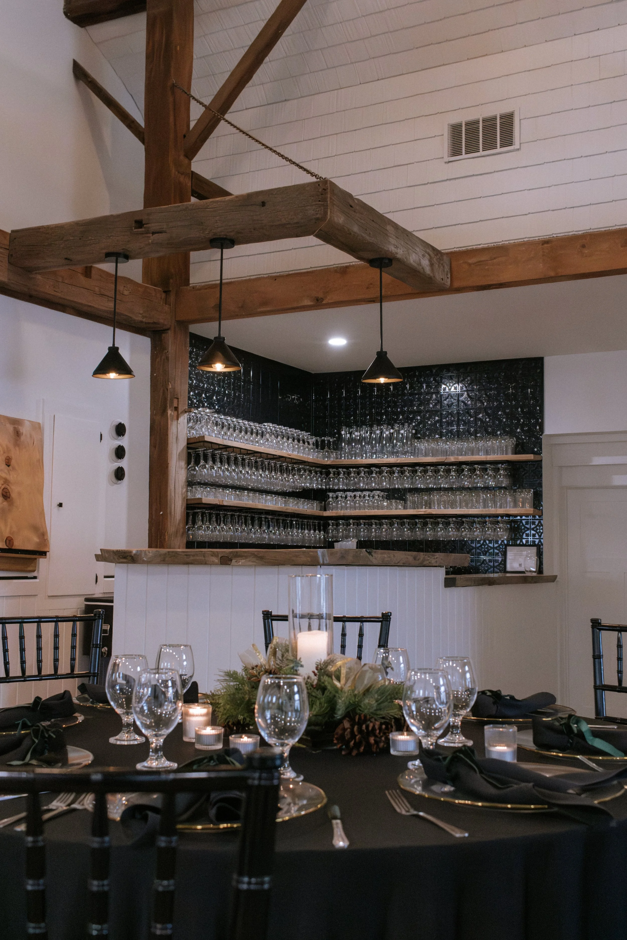 Elegant dining table set for a celebration with black tablecloth, black napkins, and glassware. A centerpiece features candles, pinecones, and greenery. A bar area in the background holds many wine glasses, with black tiles behind it and wooden shelves.