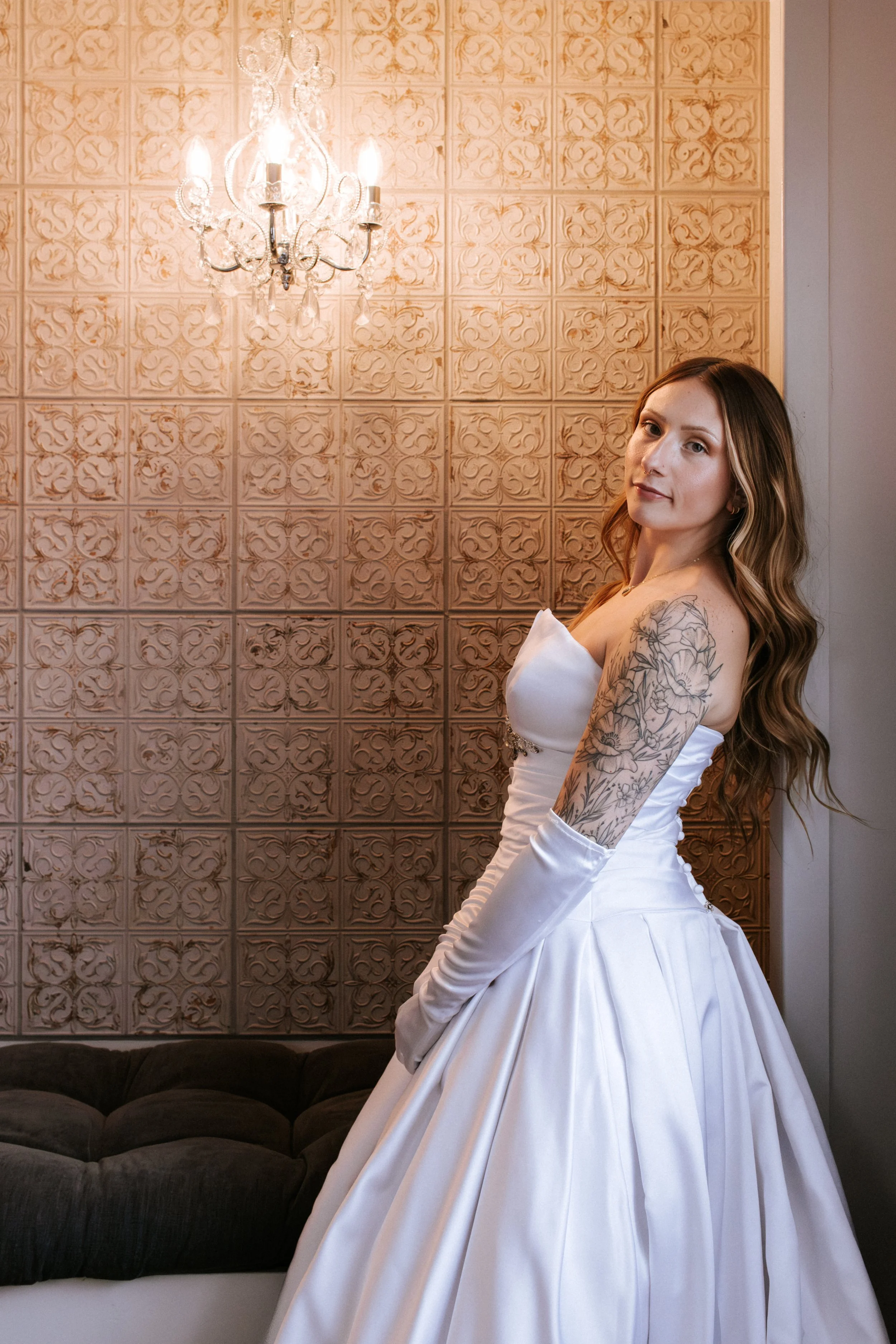 A woman in a white strapless wedding dress with long gloves standing indoors near a textured wall with a chandelier above.