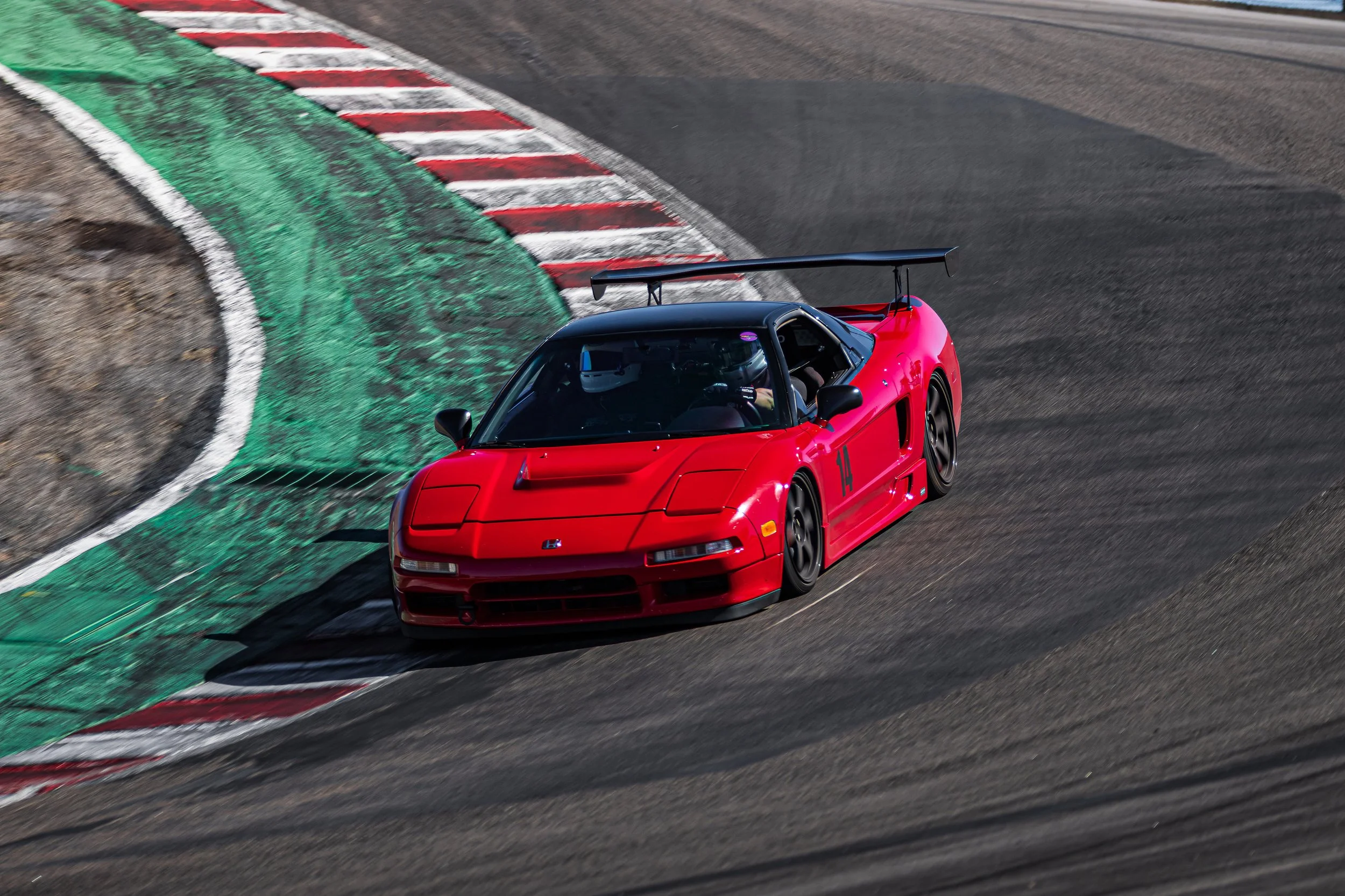 A red race car with a large rear wing is turning sharply on a racetrack with green, red, and white painted curbs.