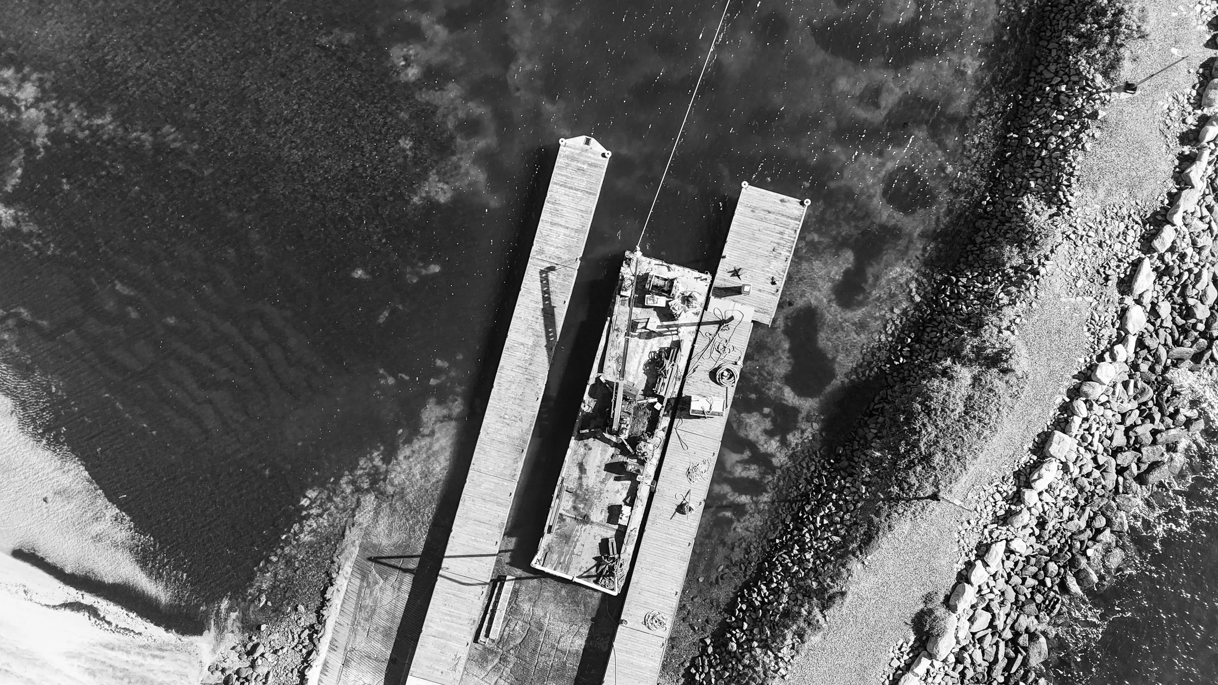 Black and white aerial view of a dock with a boat anchored, surrounded by rocks and water.