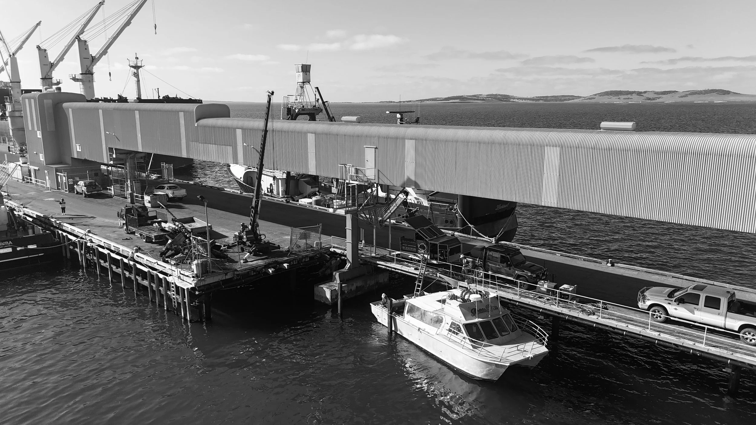 A black and white photo of a harbor with a docked boat, several cars, and a large structure above the dock, with cranes and ships in the background.