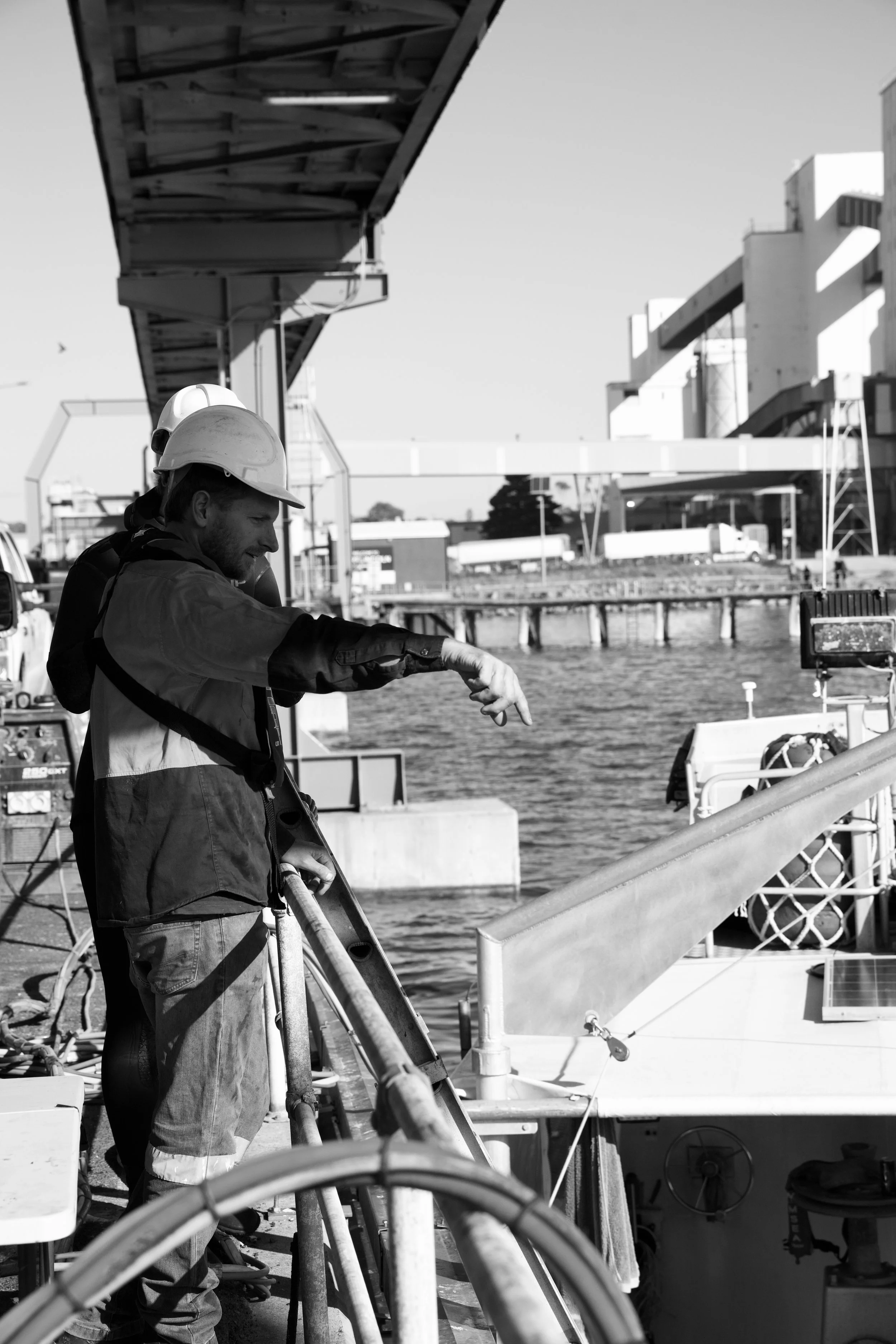 Two construction workers wearing helmets and safety gear working on a boat dock or pier by the water with industrial buildings and a bridge in the background.