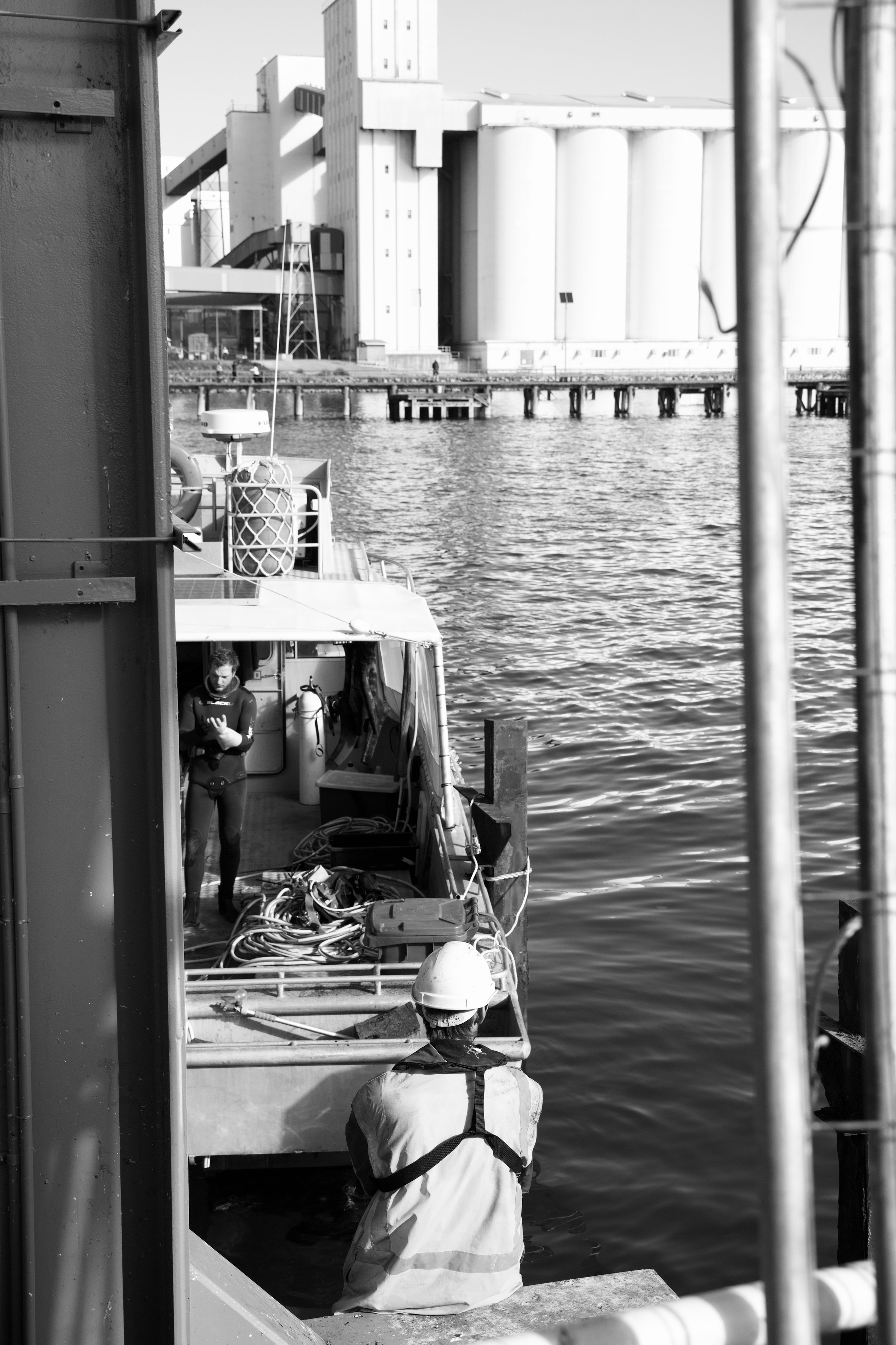 A worker with a safety helmet and vest is kneeling on the dock near a boat in a harbor, with another person standing on the boat. Industrial buildings are visible in the background.