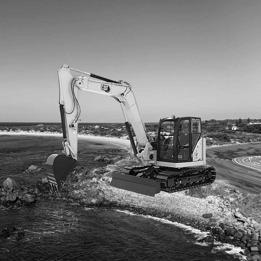 A black and white photo of a compact construction excavator with caterpillar tracks, positioned on rocky ground by a body of water, with a landscape of trees and buildings in the background.