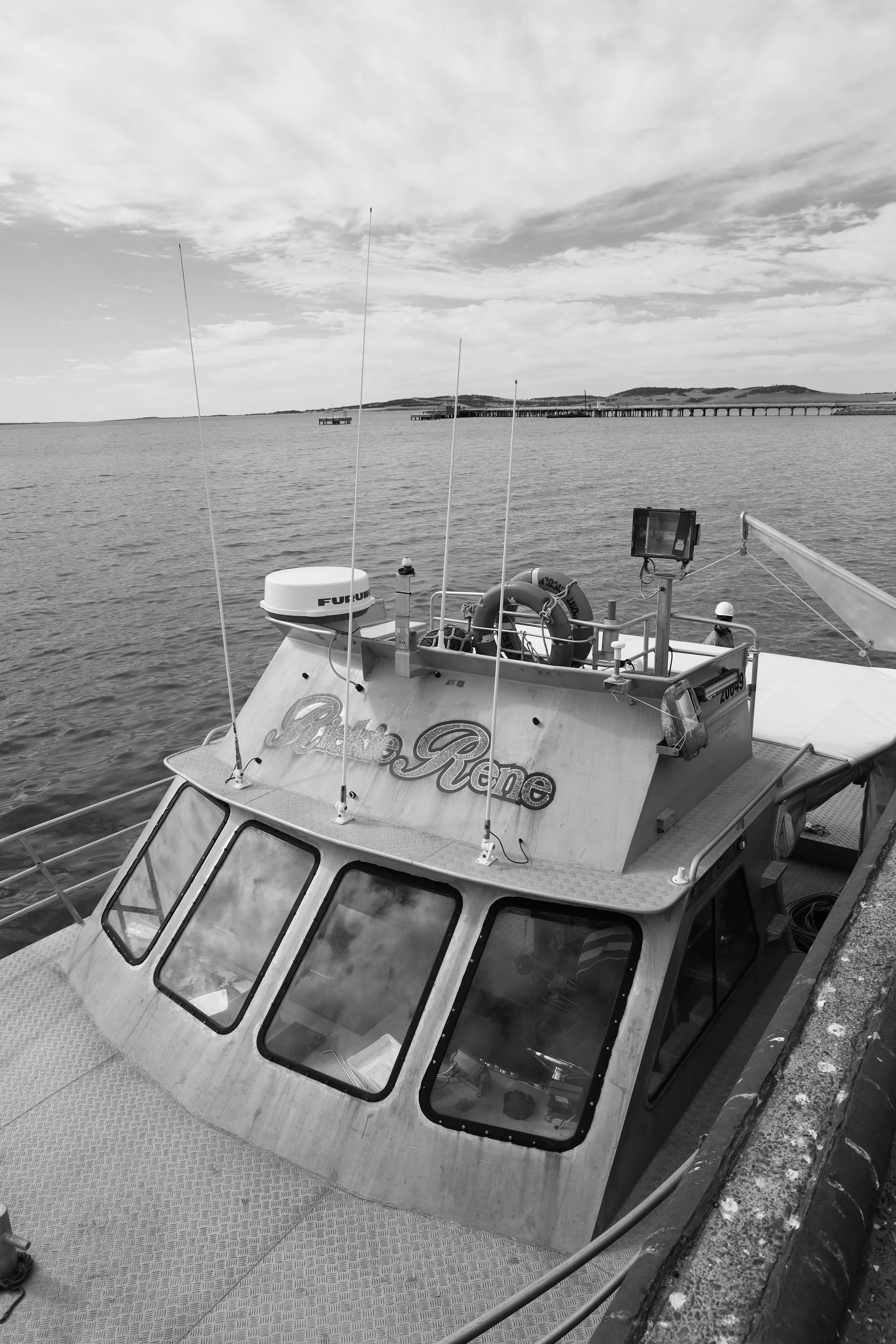 A boat docked at a pier on a calm body of water, with a distant shoreline and cloudy sky in the background.