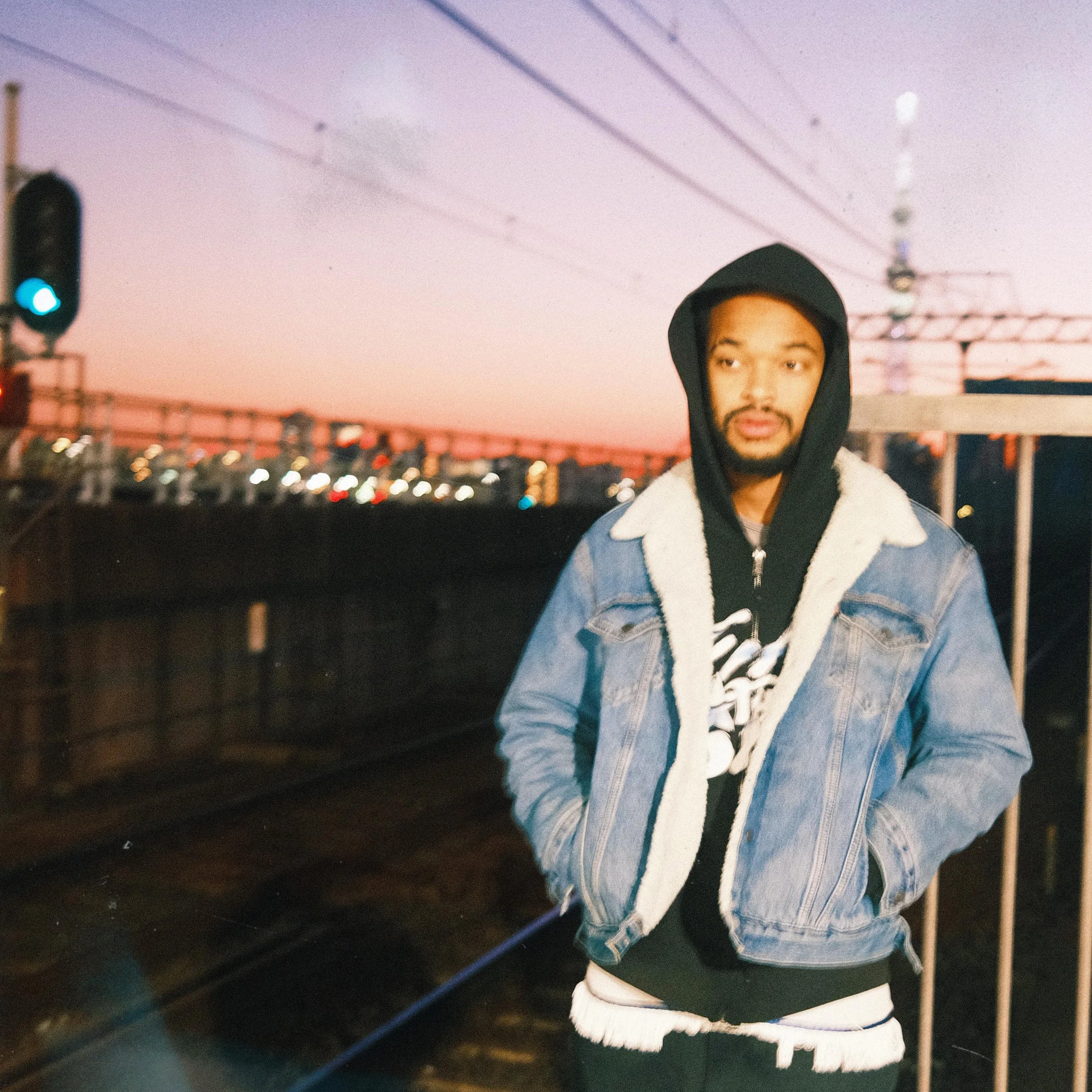 Jx.Zero wearing a black hoodie and a denim jacket standing on a train platform at sunset.
