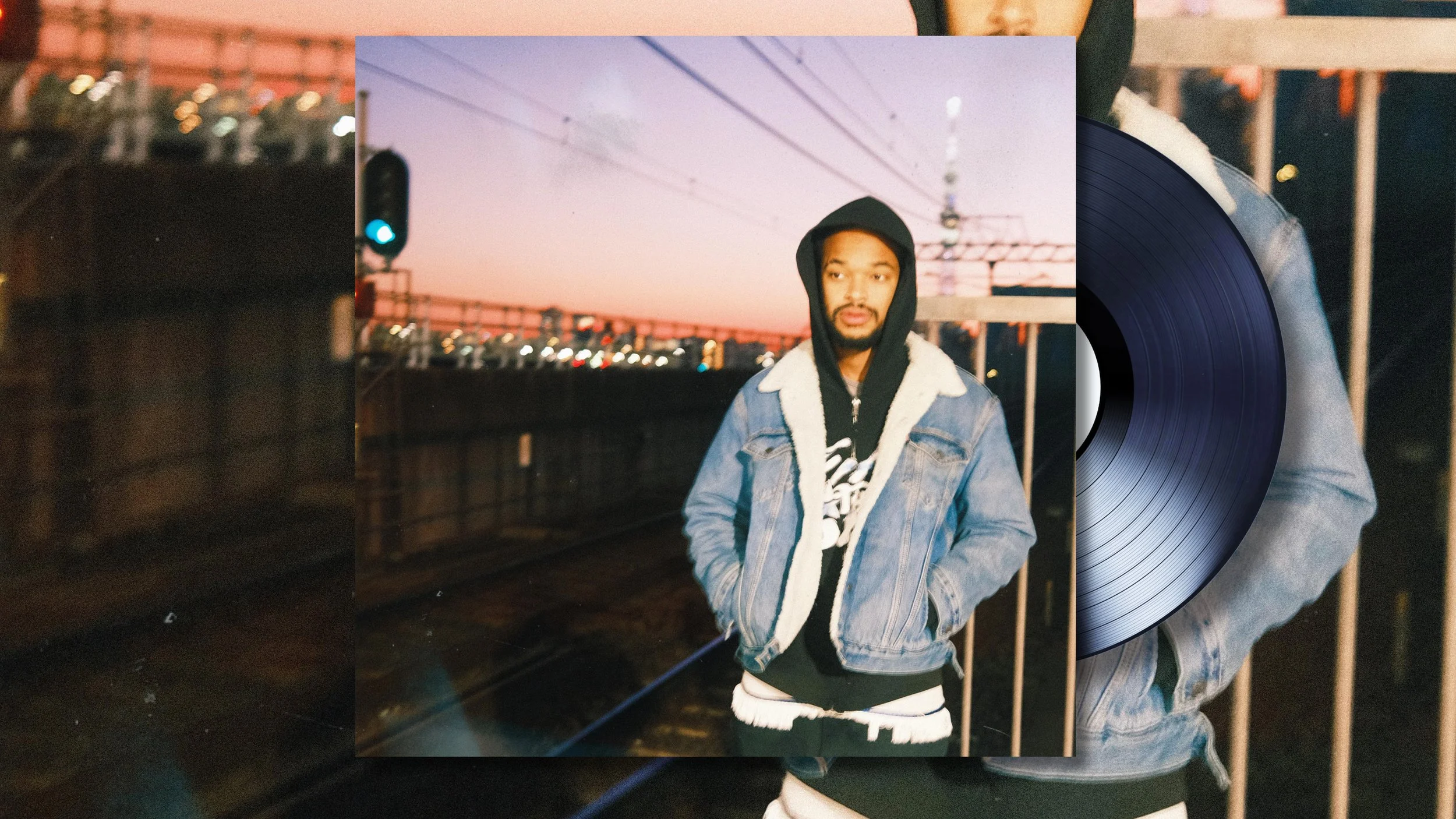 Jx.Zero wearing a black hoodie, denim jacket with a shearling collar, and dark pants standing outdoors during sunset near train tracks, with an overpass and city lights in the background.