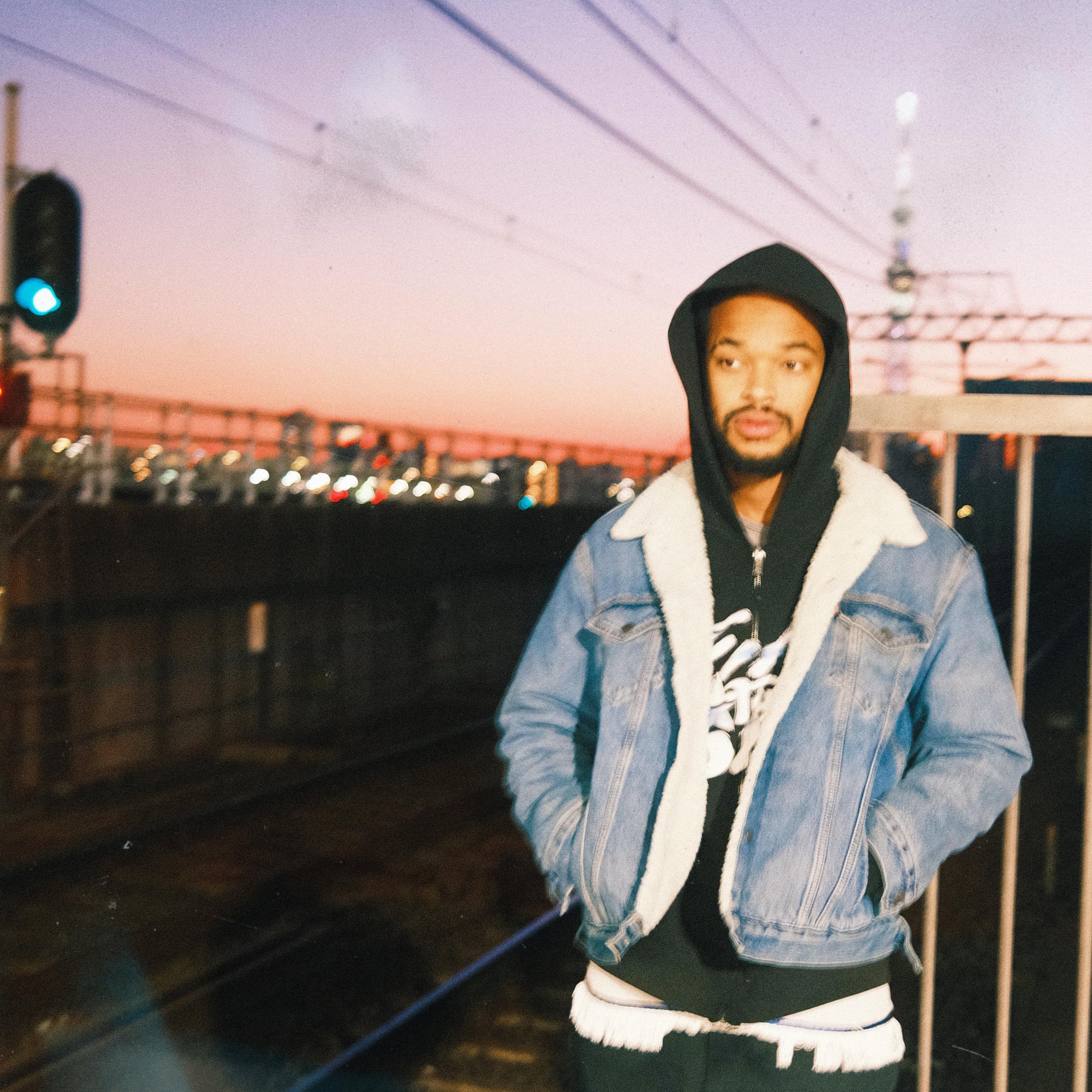Jx.Zero wearing a black hoodie, denim jacket with a shearling collar, and dark pants standing outdoors during sunset near train tracks, with an overpass and city lights in the background.