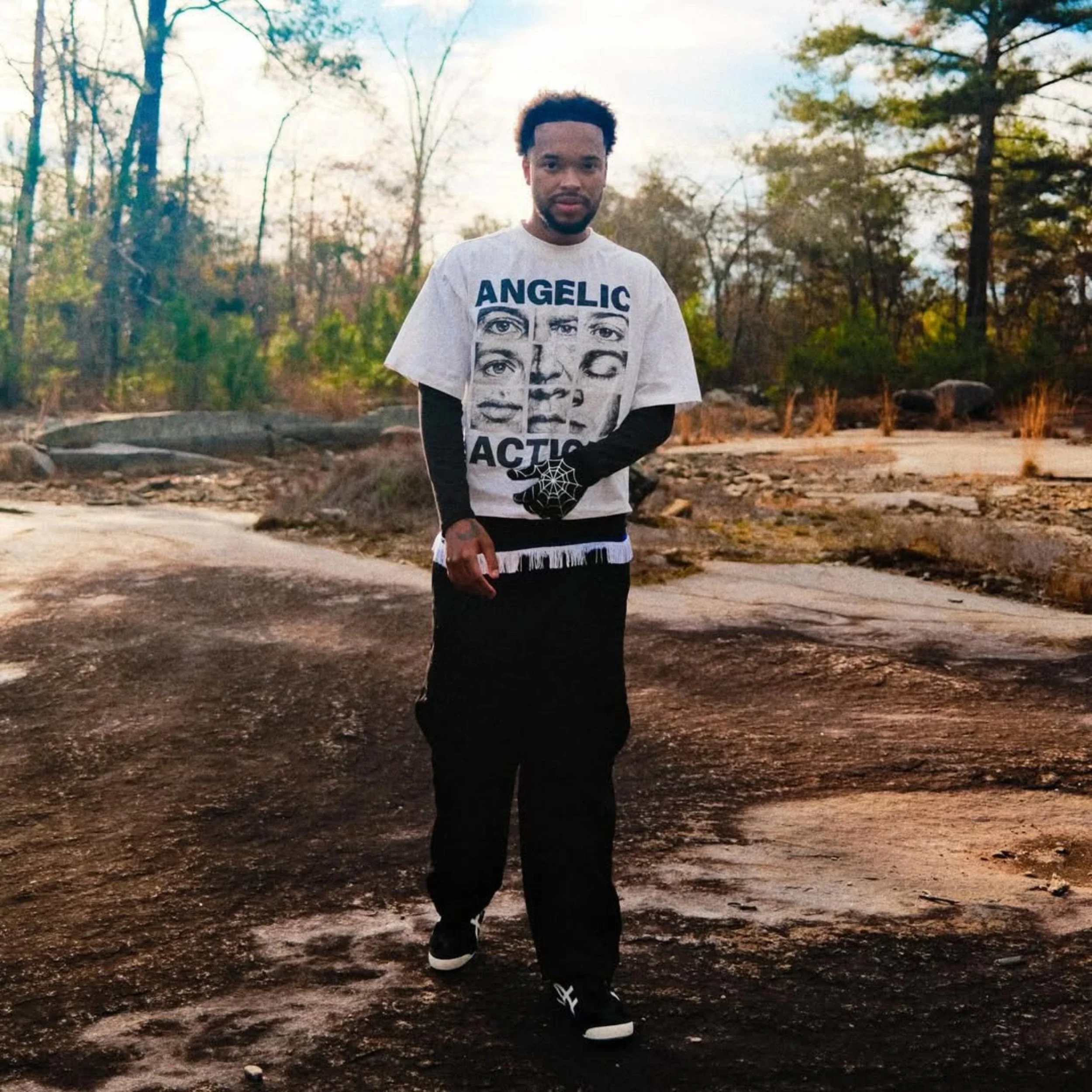 Jx.Zero standing outdoors on a rocky path surrounded by trees with autumn foliage, wearing a graphic T-shirt and black pants.