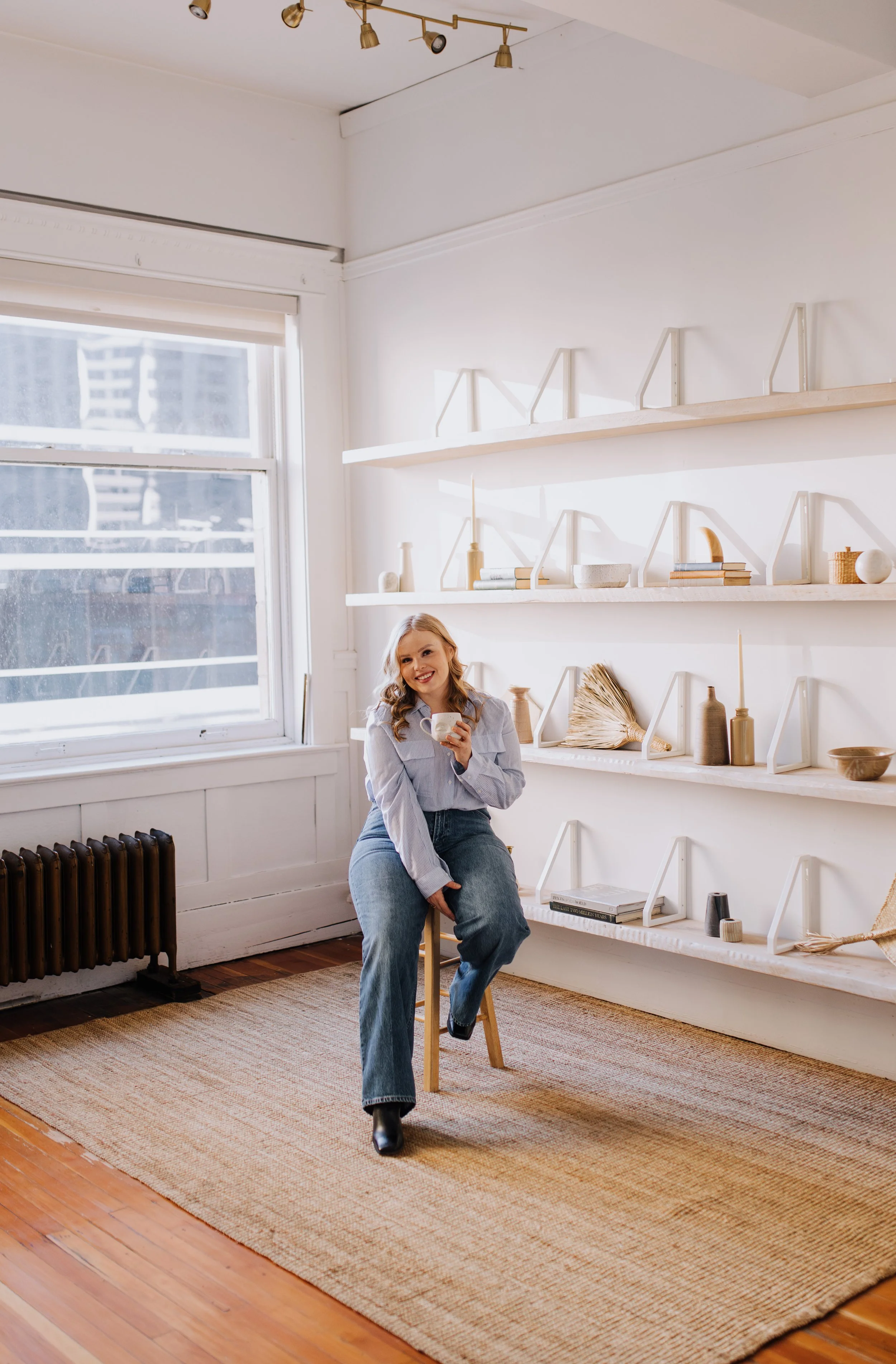 A woman sitting on a wooden chair in a bright room, holding a mug and smiling. The room has white walls, shelves with decorative items, a large window with sunlight, a radiator, and a woven rug on a wooden floor.