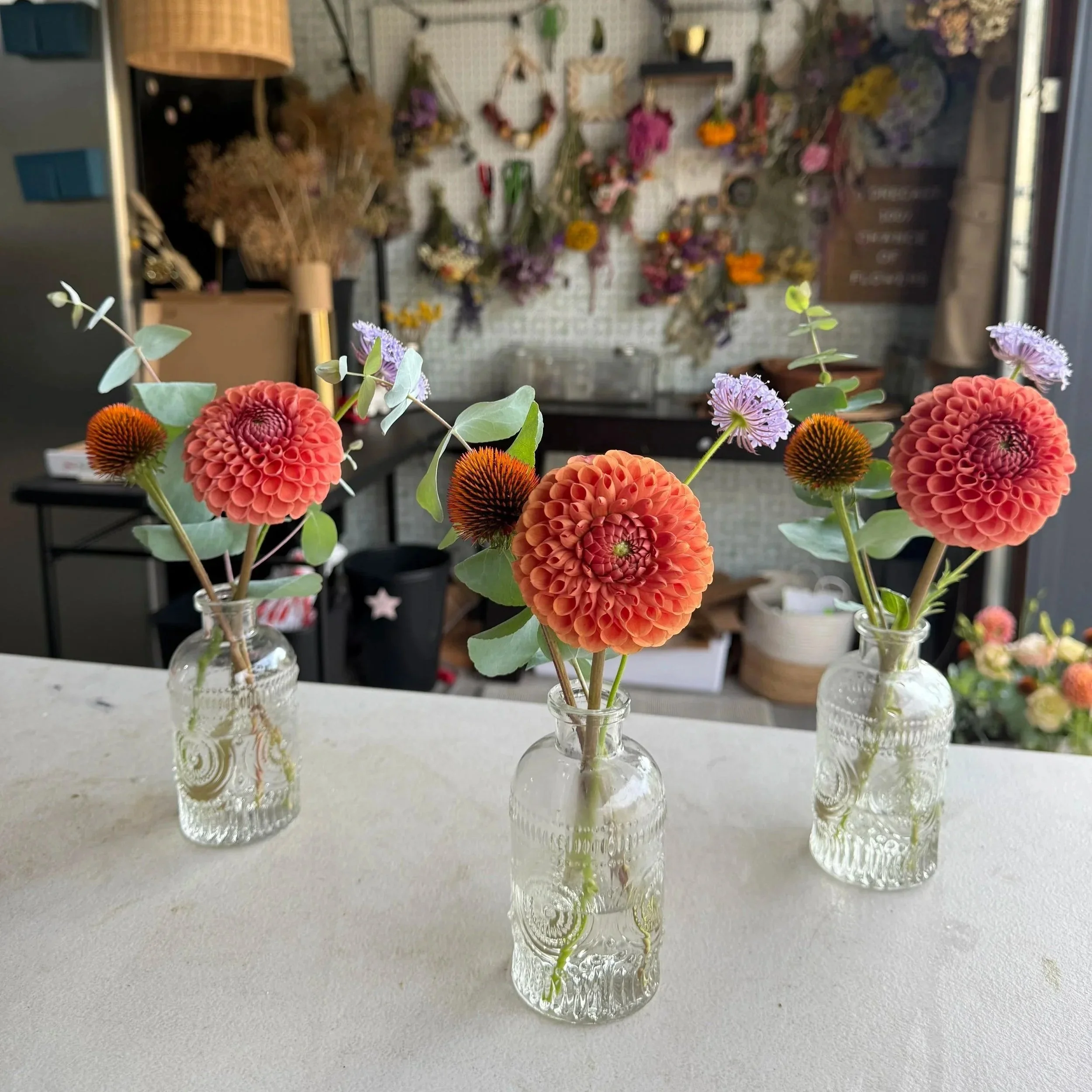Three small glass vases with dried flowers on a white surface, with a floral shop in the background.