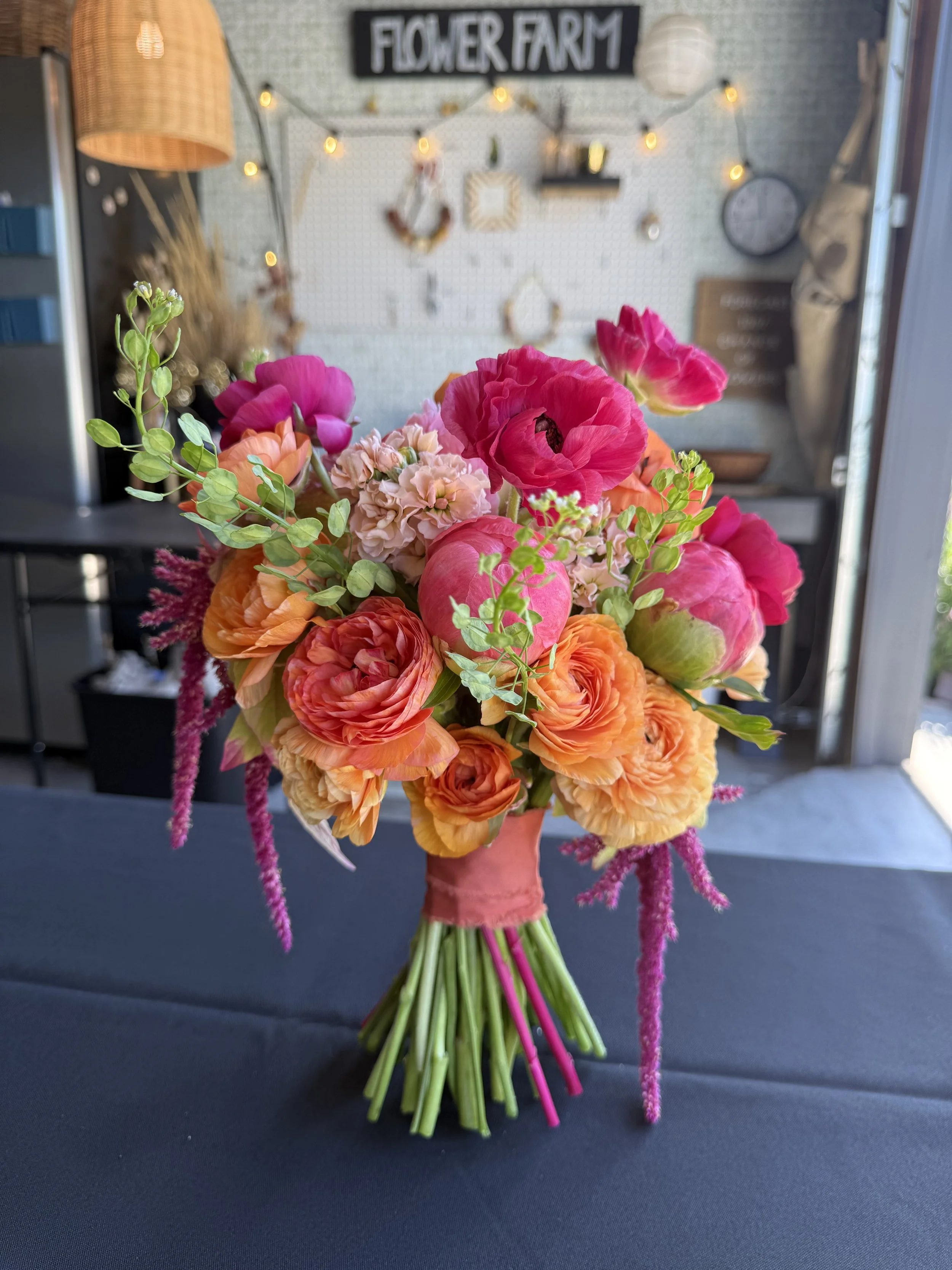 A colorful bouquet of pink, orange, peach, and purple flowers with green leaves, arranged in a pink wrapping, placed on a dark surface with a blurred background of a sign reading 'FLOWER FARM' and string lights.