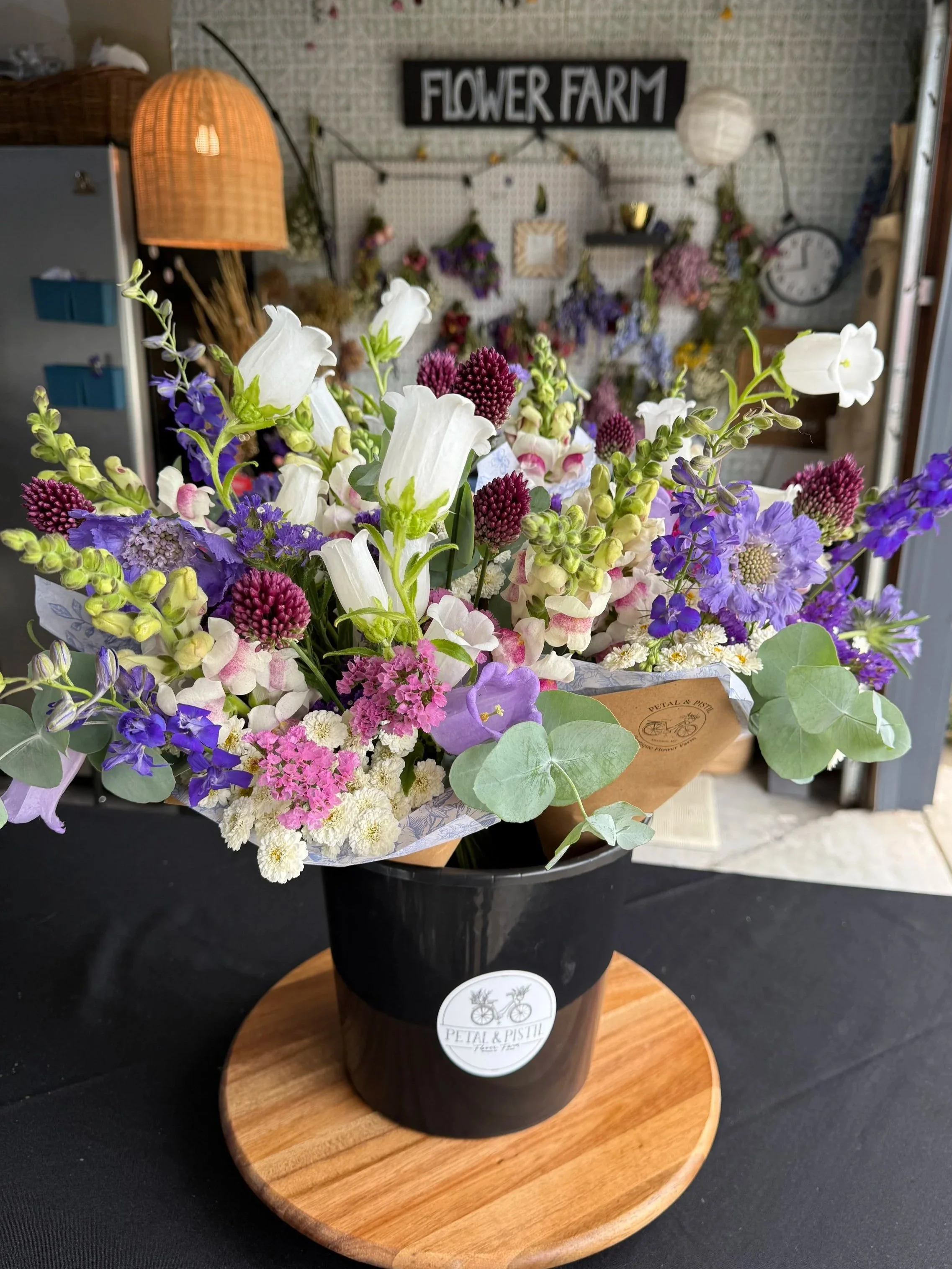 A bouquet of mixed flowers in a black pot with a wooden base, placed on a round wooden tray, with a blurred background including a sign reading 'FLOWER FARM'.