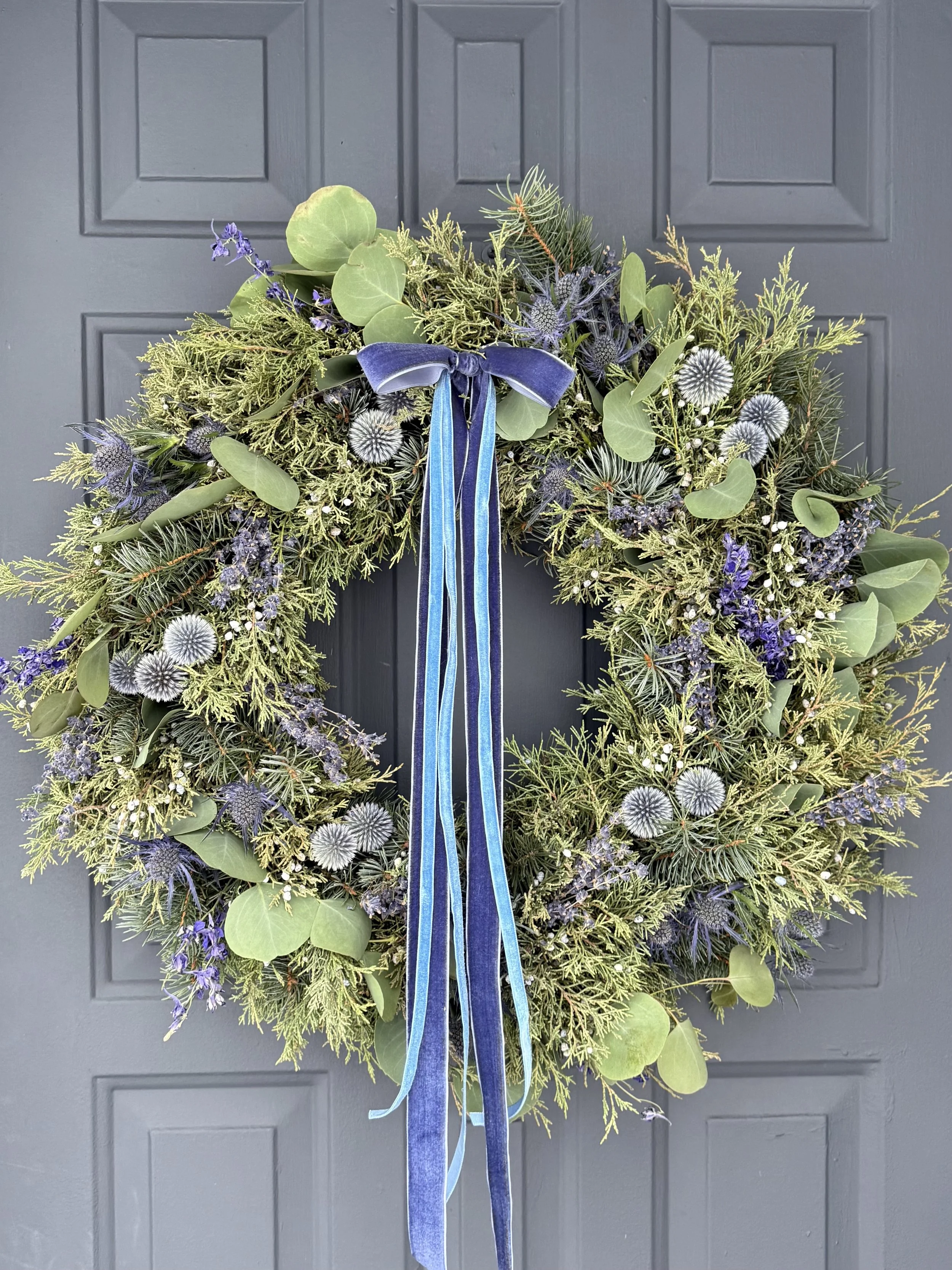 A wreath made of greenery, purple and white flowers, and a blue ribbon, hanging on a gray door.