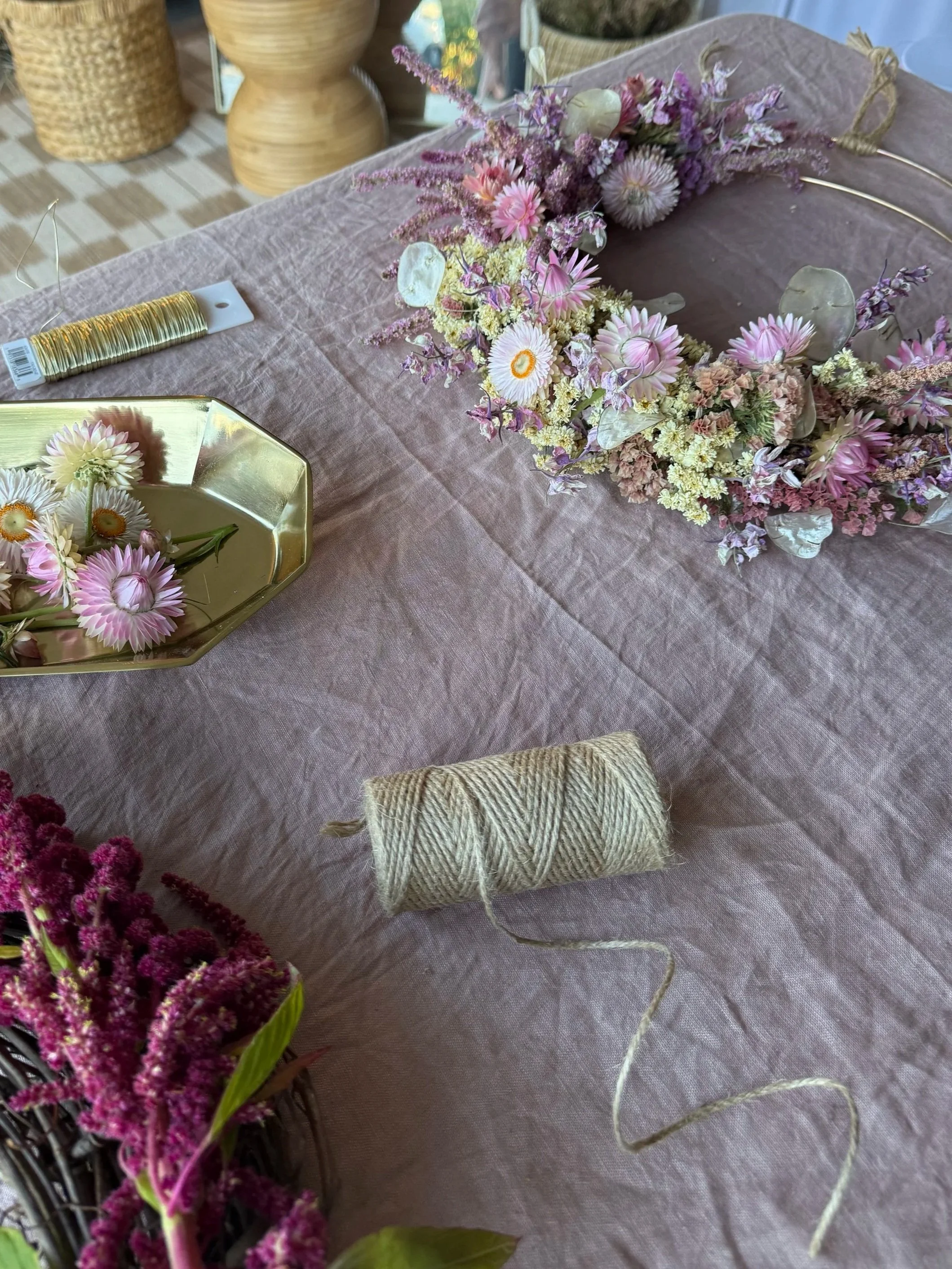 A flower arrangement with pink and purple flowers on a table covered with pink fabric, alongside a spool of twine, a gold tray with pink and white flowers, and wooden decorative objects in the background.