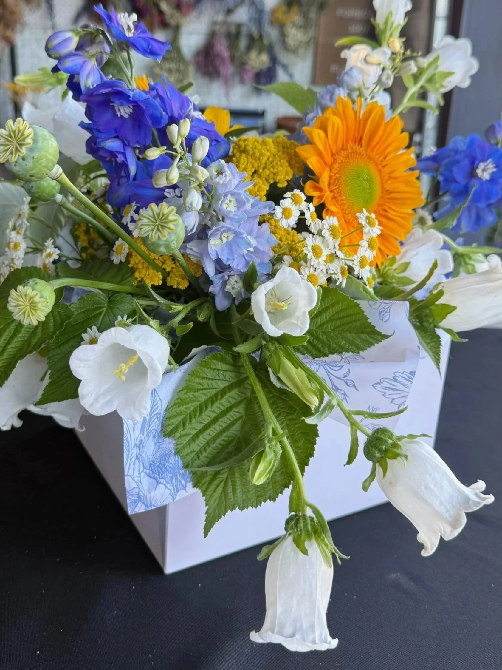 A colorful bouquet of blue, yellow, orange, and white flowers wrapped in white paper, resting on a black surface.